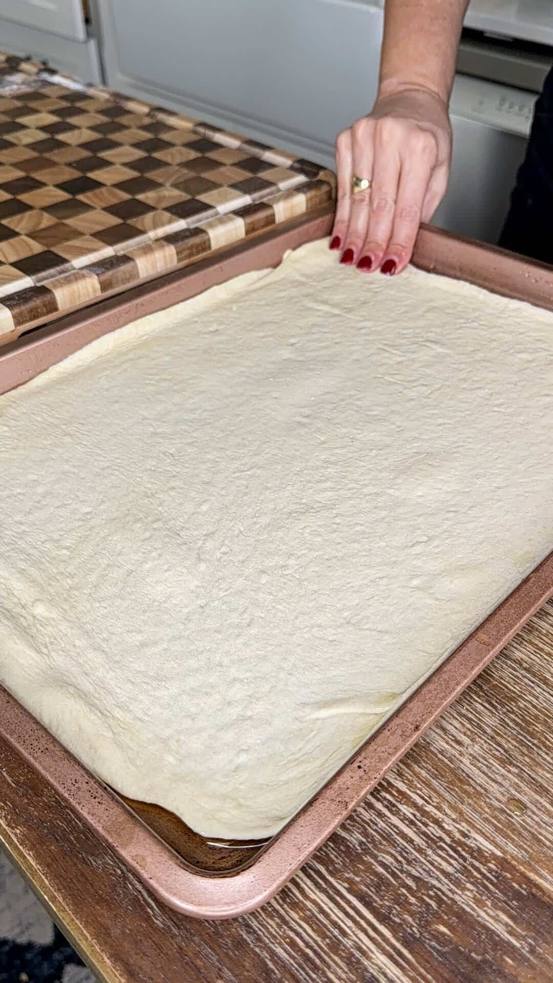 A persons hand stretches a sheet of raw pizza dough to fit into a rectangular copper-colored baking pan.