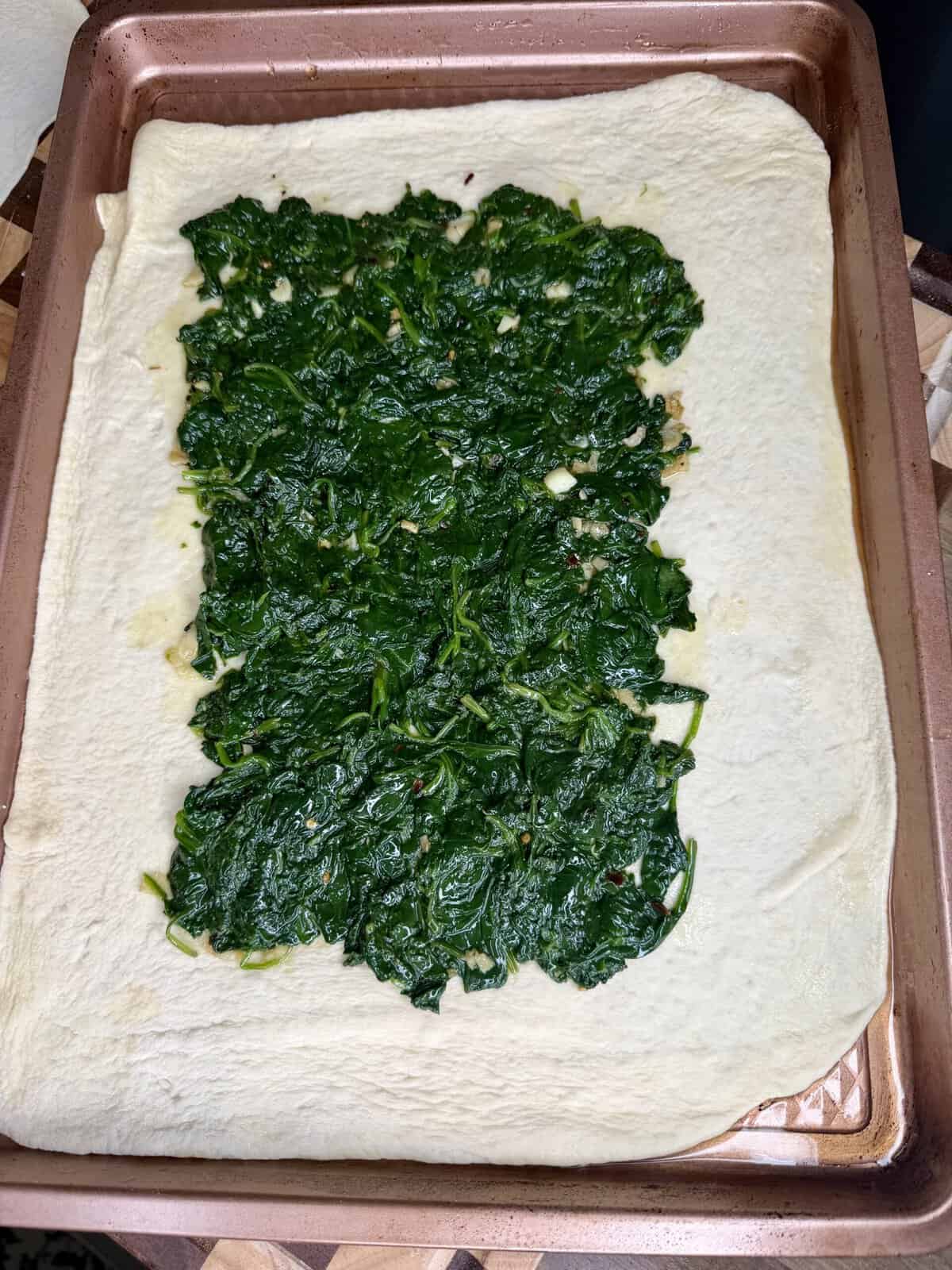 A sheet of raw dough on a baking pan topped with a rectangular layer of cooked, dark green spinach.