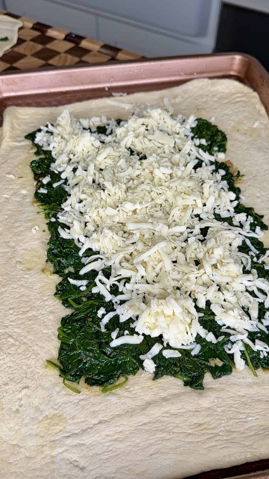 A rectangular sheet of raw dough topped with a layer of cooked spinach and shredded cheese.