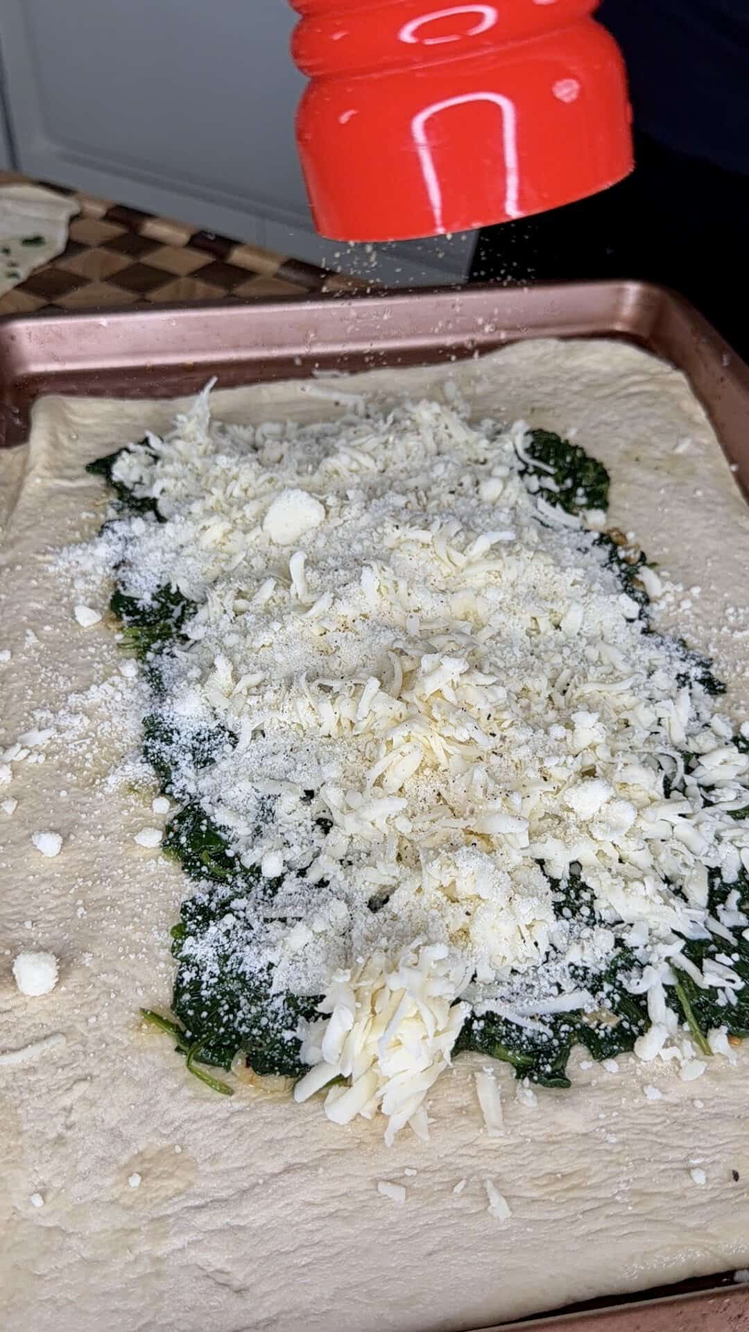 A red grater sprinkles grated cheese over a layer of spinach on top of rolled-out pizza dough in a baking pan.