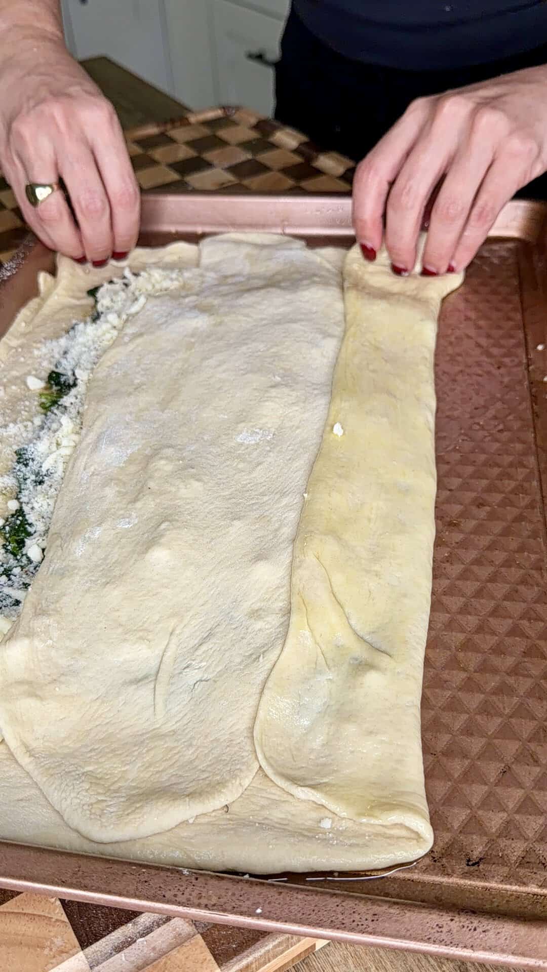Hands folding a sheet of dough over a spinach and cheese filling on a baking sheet.