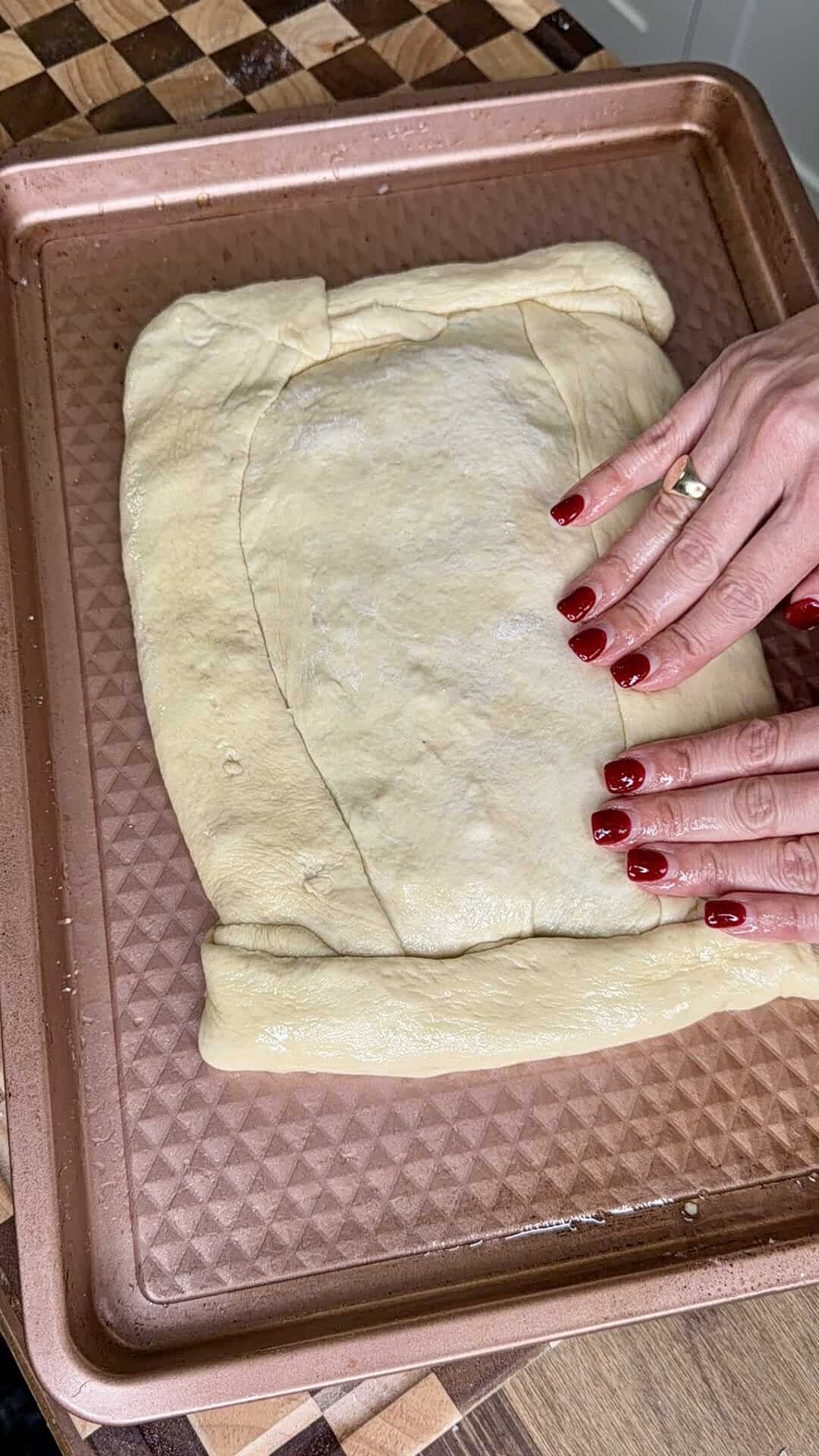 Hands with red nail polish press the edges of a rectangular piece of raw dough on a copper-colored baking sheet.