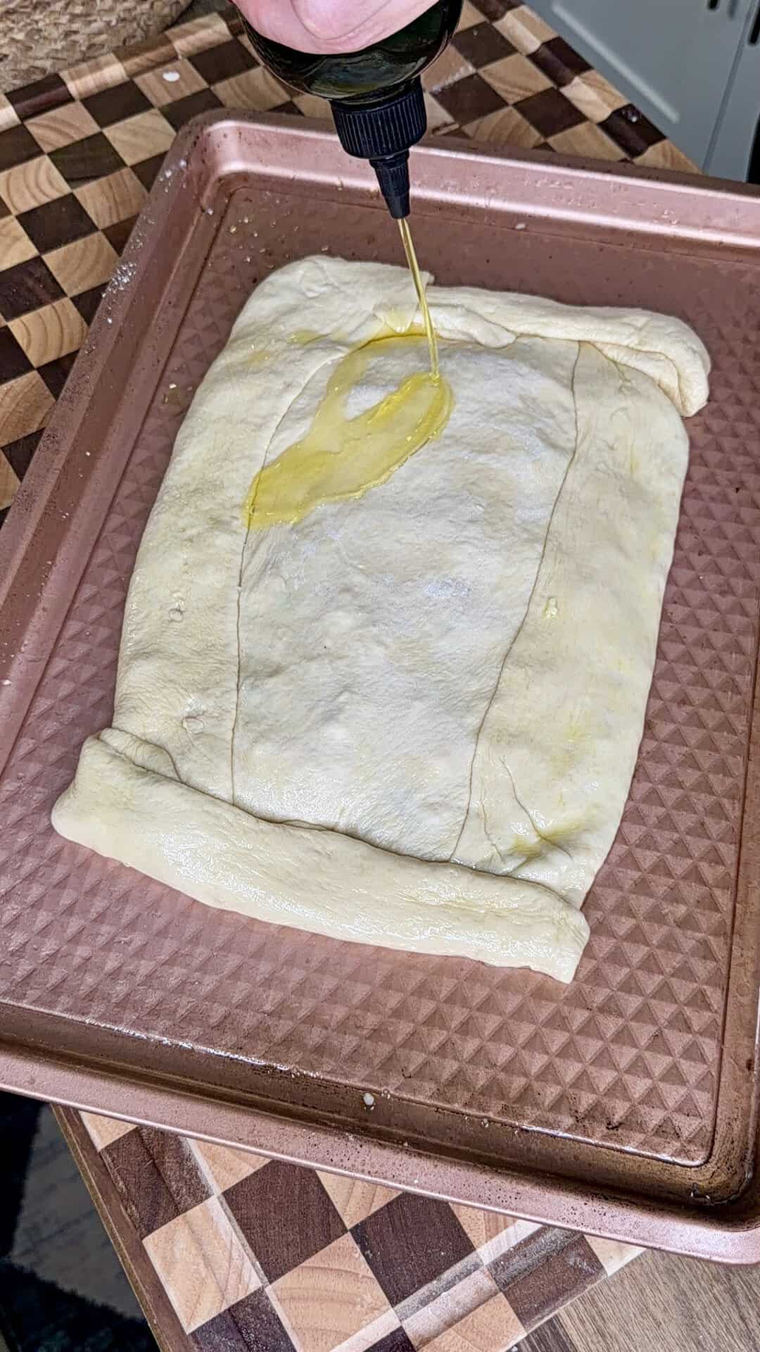 Olive oil being drizzled from a squeeze bottle onto a folded rectangle of raw dough on a baking sheet.