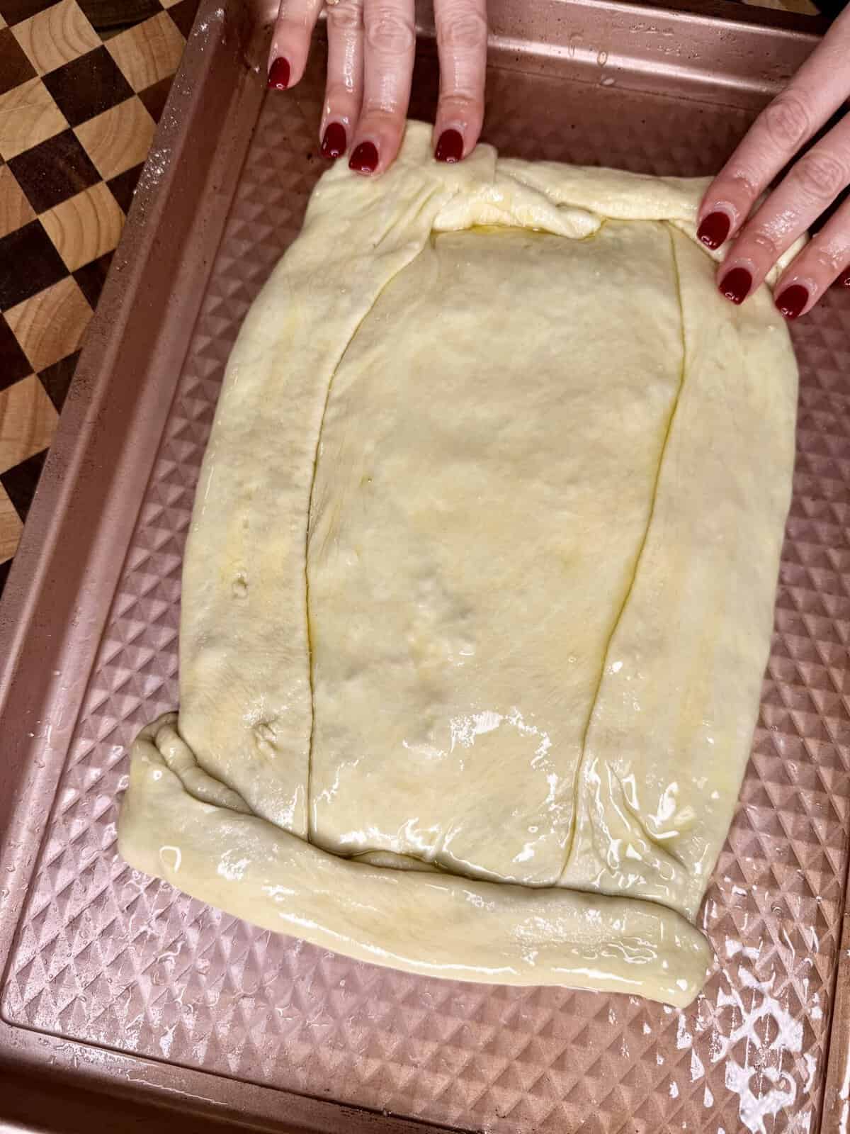 Hands fold the edges of a rectangular piece of raw dough on a baking sheet.