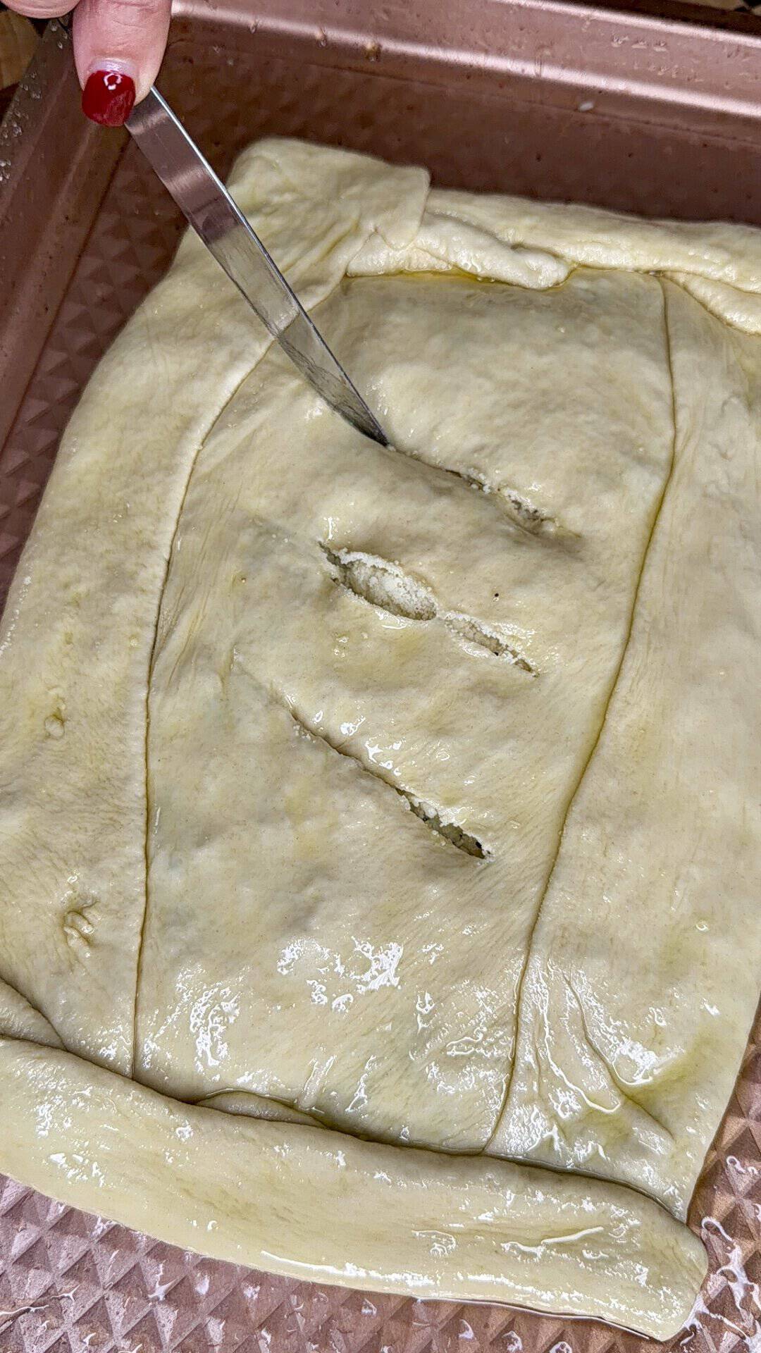 A hand with red nail polish uses a knife to cut steam vents into a rectangular piece of unbaked dough in a baking pan.
