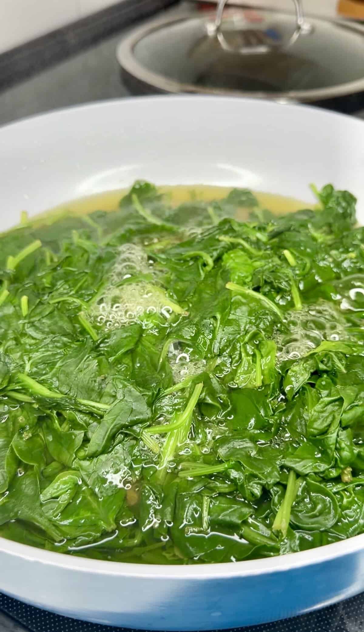 Fresh spinach leaves simmering in a shallow pan of liquid.