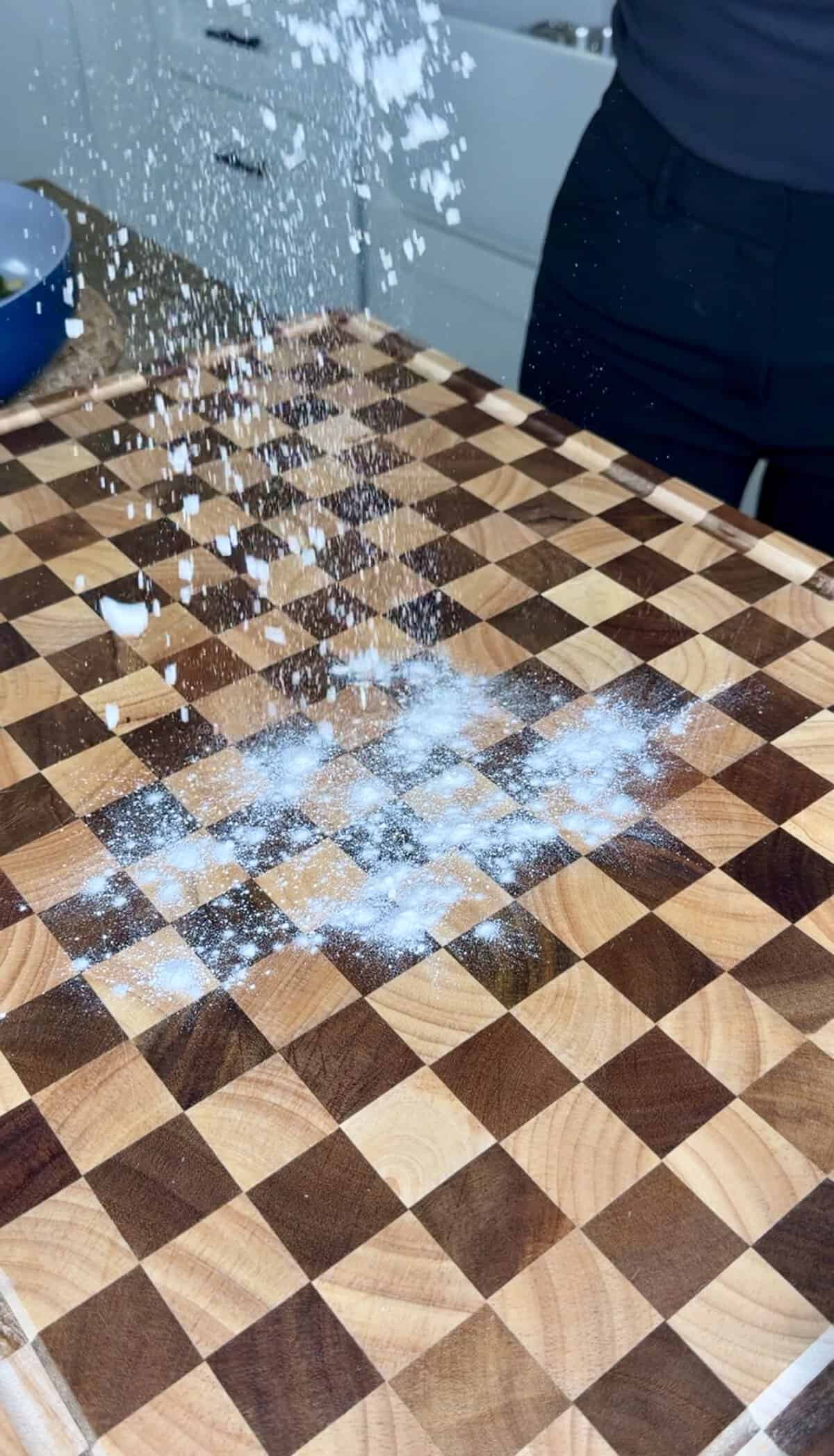 Flour being dusted onto a checkerboard-patterned wooden cutting board.