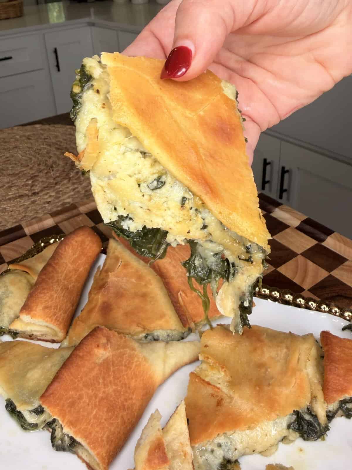 A hand holds a triangular slice of golden-brown spinach and cheese pie, with more pieces resting on a plate below.