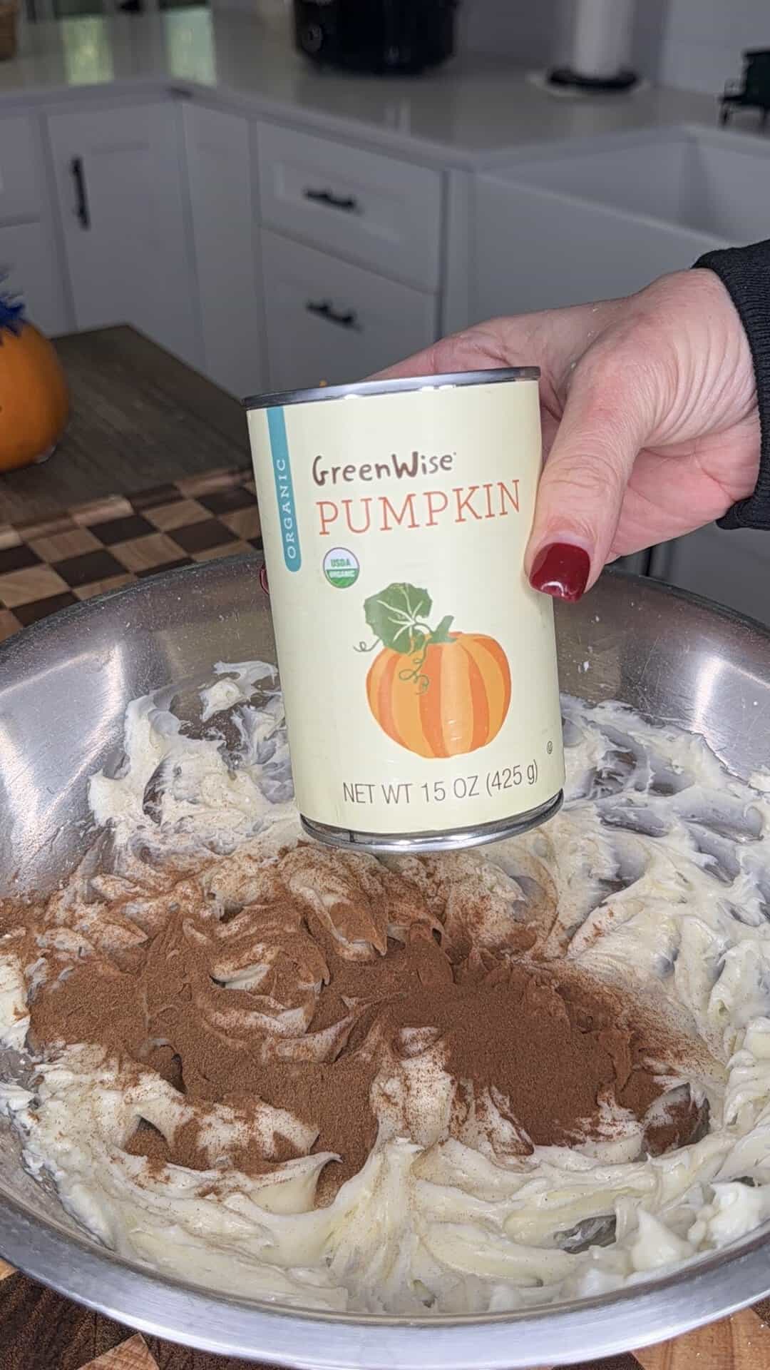 A hand holds a can of GreenWise organic pumpkin over a metal bowl containing creamed ingredients topped with ground spices.