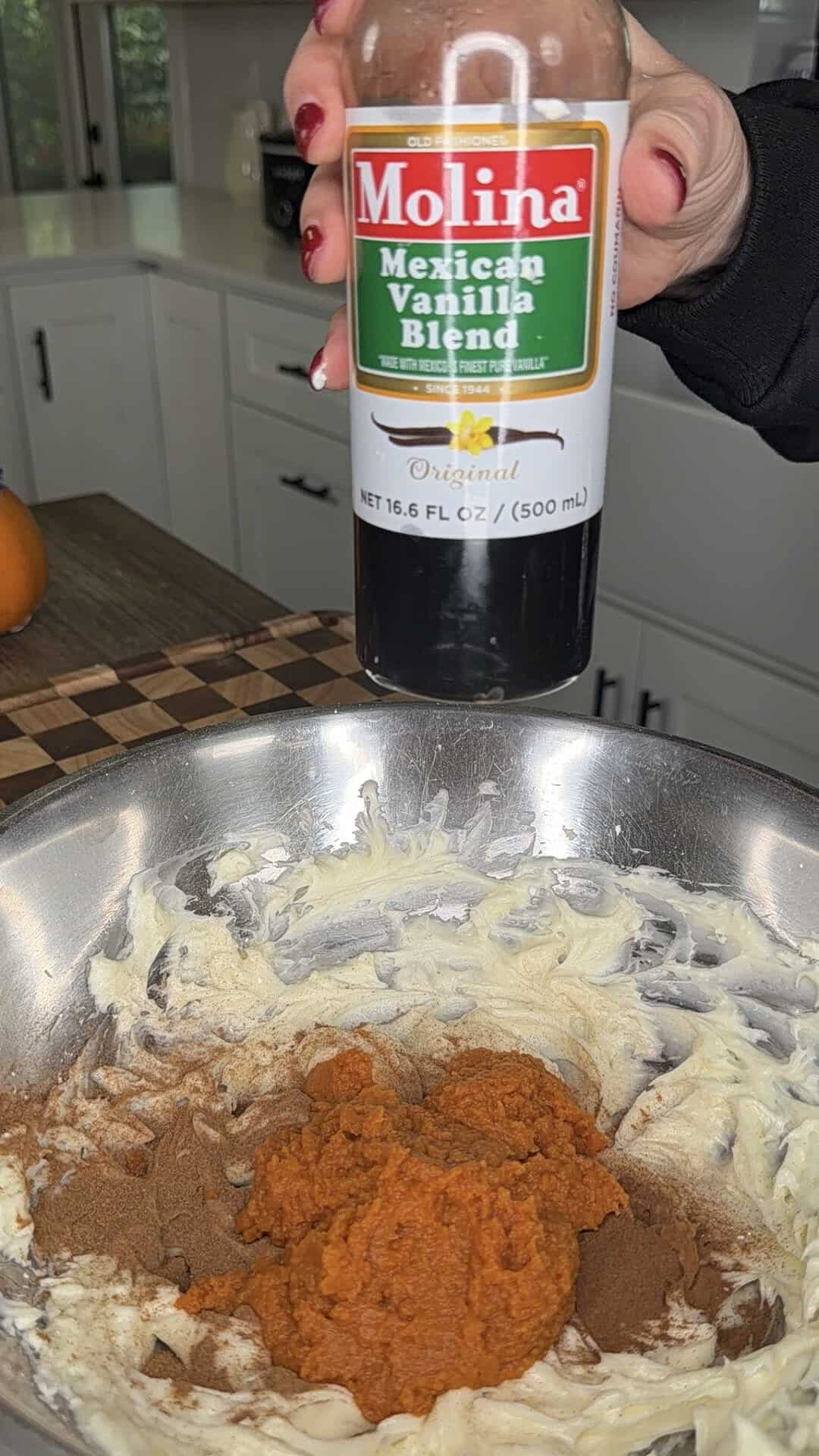 A hand holds a bottle of Molina Mexican Vanilla Blend over a metal mixing bowl containing creamed butter, pumpkin puree, and spices.