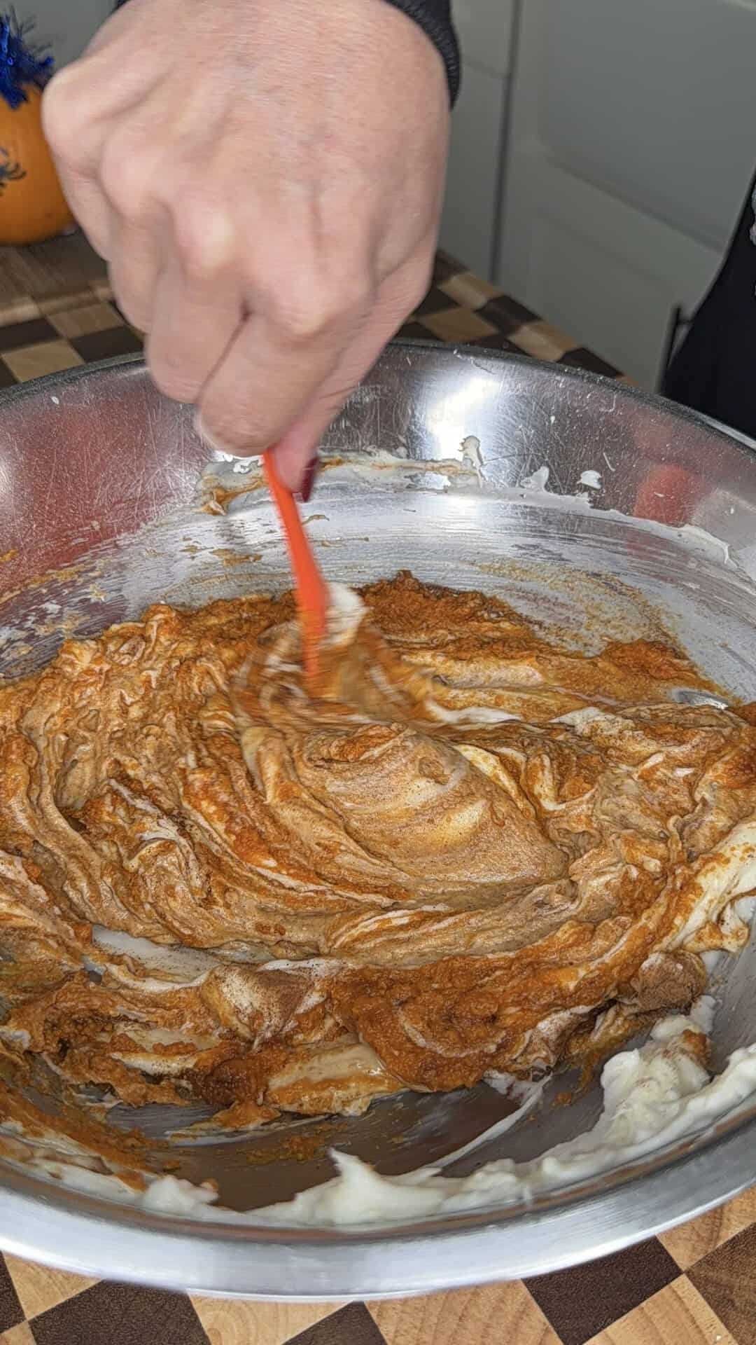 A hand uses a red spatula to fold pumpkin batter into white cream in a metal mixing bowl.