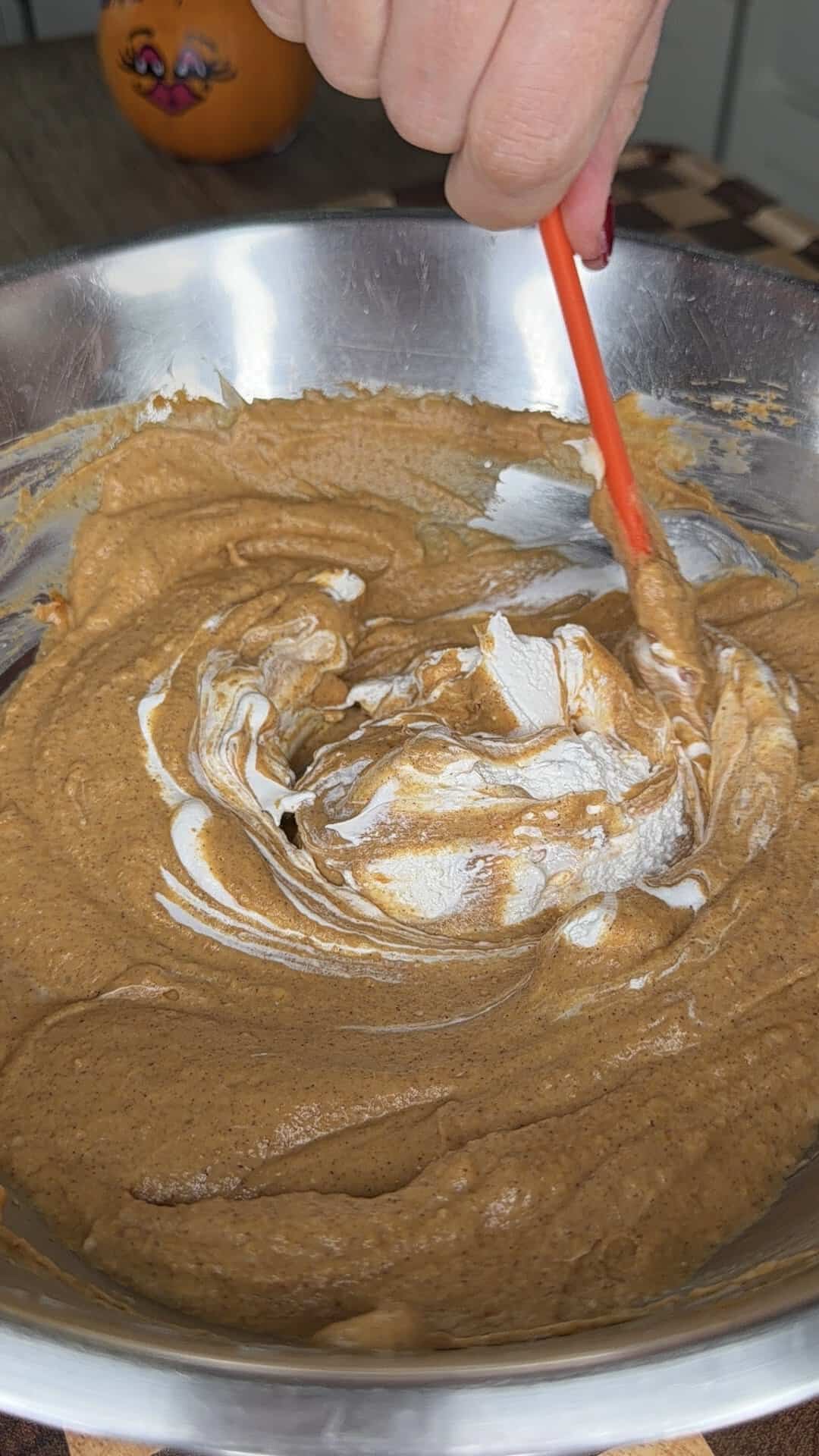 A hand uses an orange spatula to fold white cream into a bowl of brown batter.