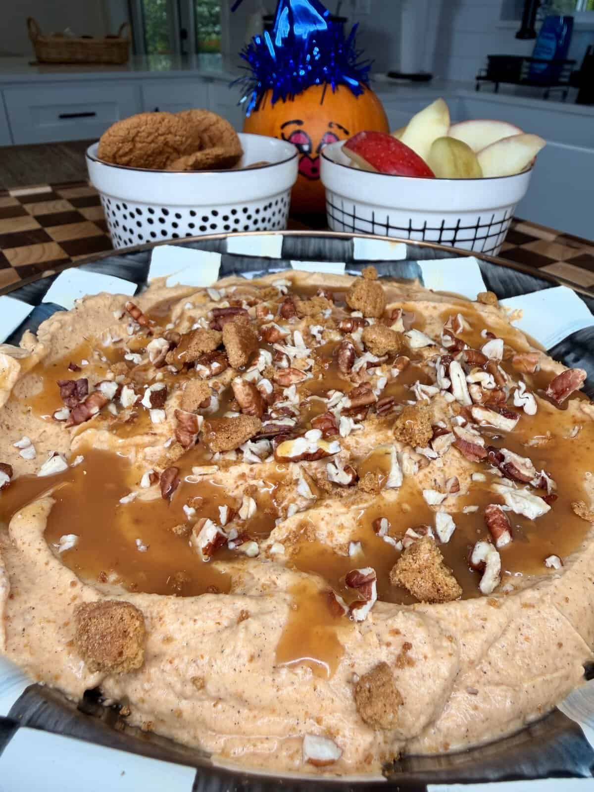 A bowl of pumpkin dip topped with caramel sauce, chopped pecans, and cookie crumbles, served with side bowls of apple slices and cookies.