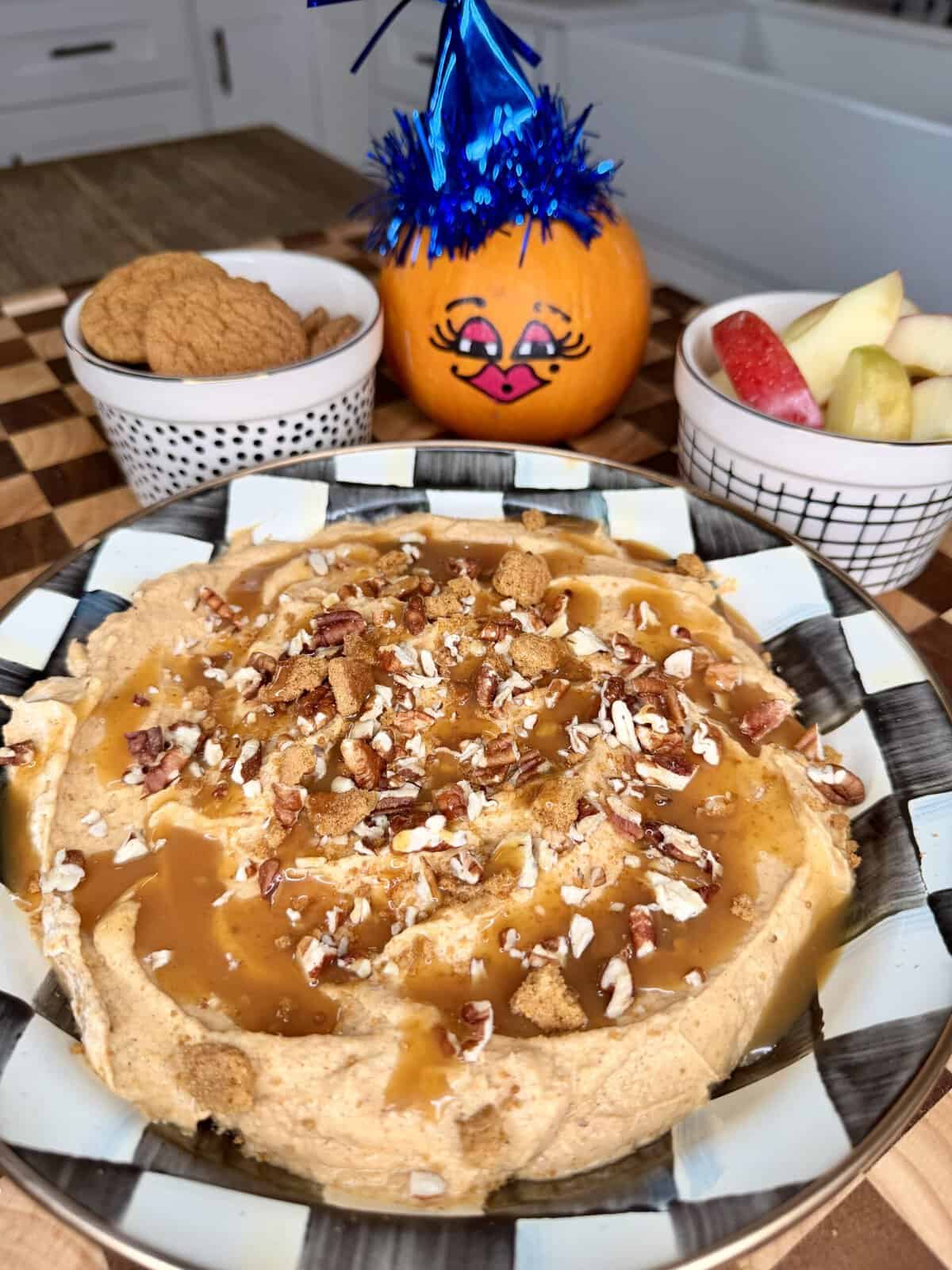 A bowl of caramel-drizzled pumpkin dip topped with pecans and cookie crumbles, served with bowls of ginger cookies and sliced apples beside a small painted pumpkin.