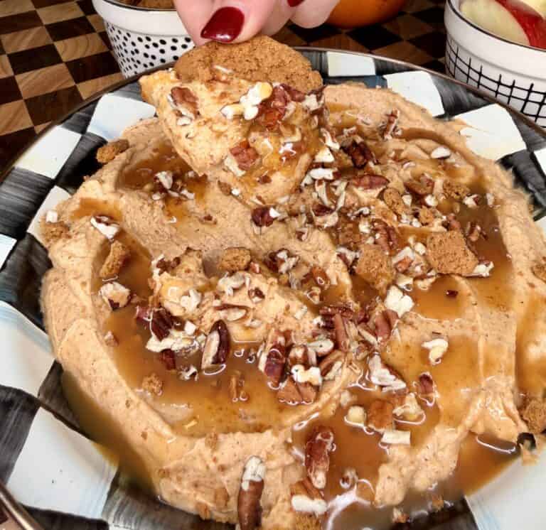 A hand dips a gingersnap cookie into a bowl of pumpkin dip drizzled with caramel and topped with chopped pecans.