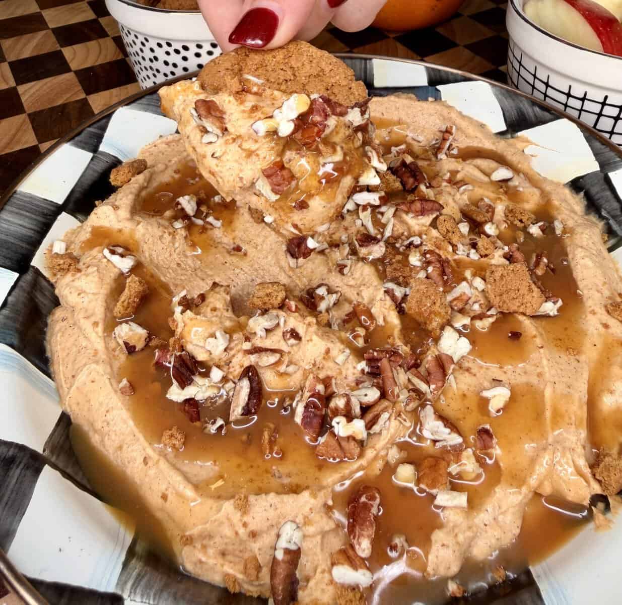 A hand dips a gingersnap cookie into a bowl of pumpkin dip drizzled with caramel and topped with chopped pecans.