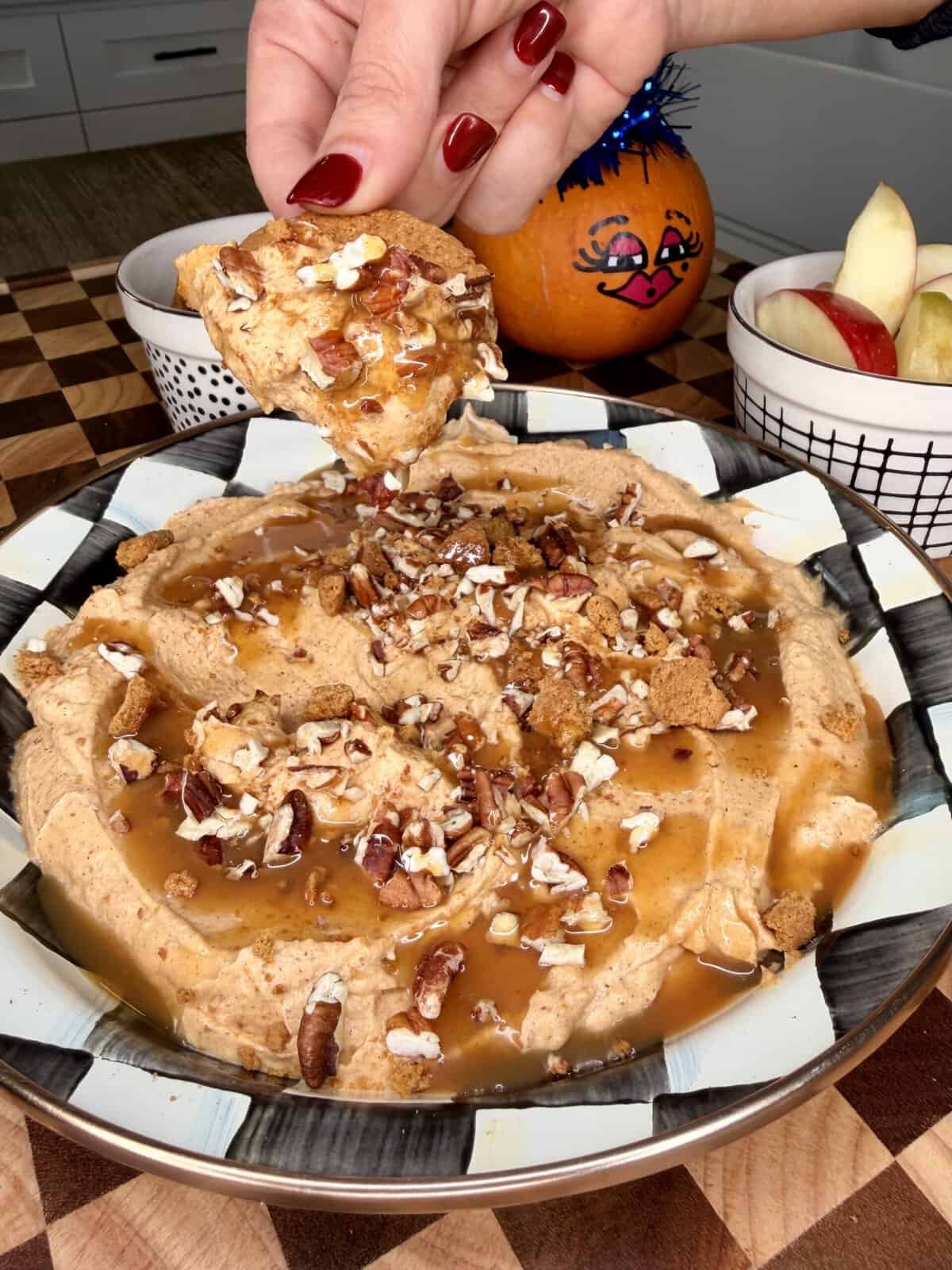 A hand dips a graham cracker into a bowl of pumpkin pie dip topped with caramel sauce and chopped pecans.