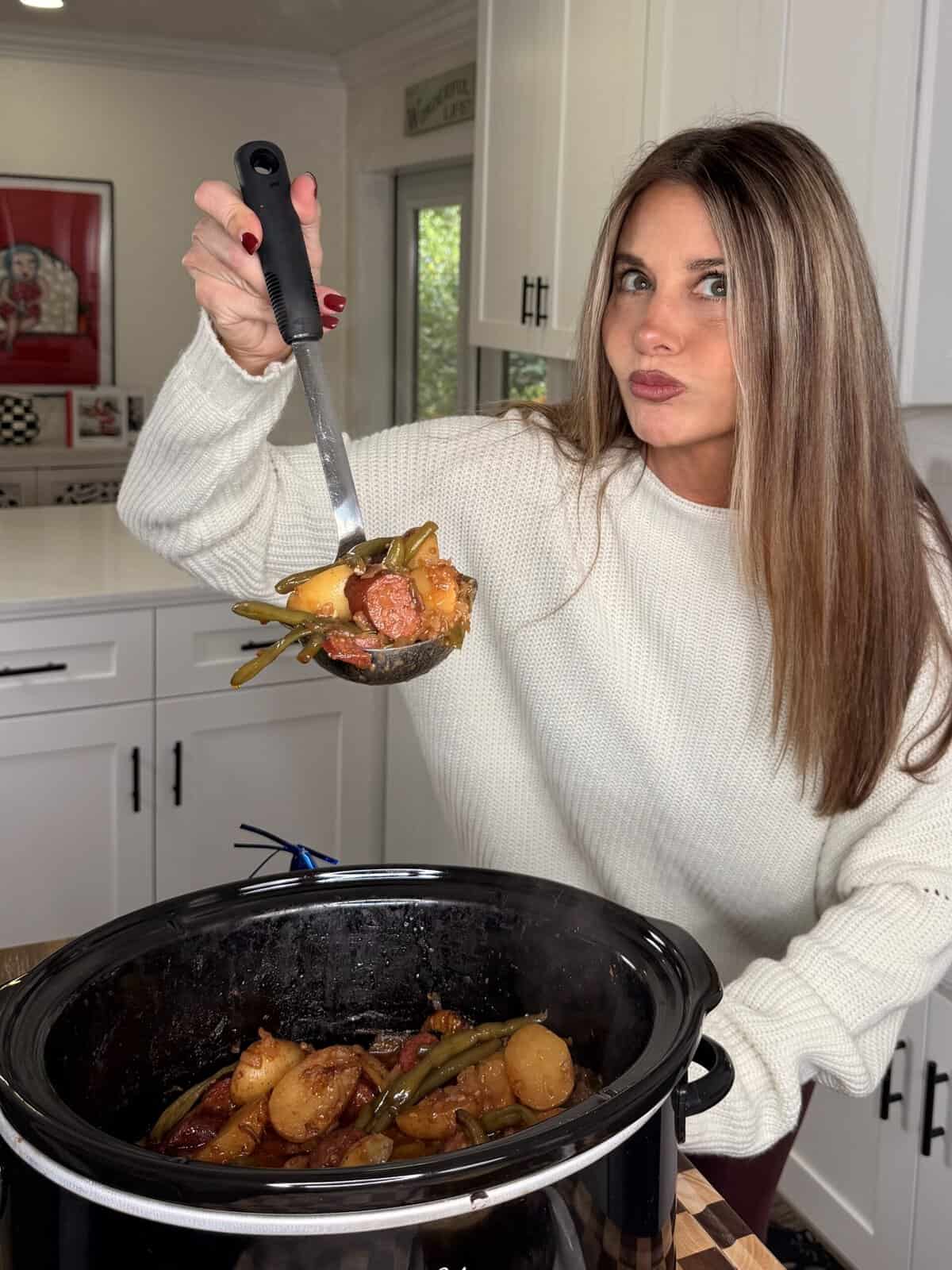 A woman in a white sweater holds a ladle full of cooked sausage, potatoes, and green beans above a slow cooker.