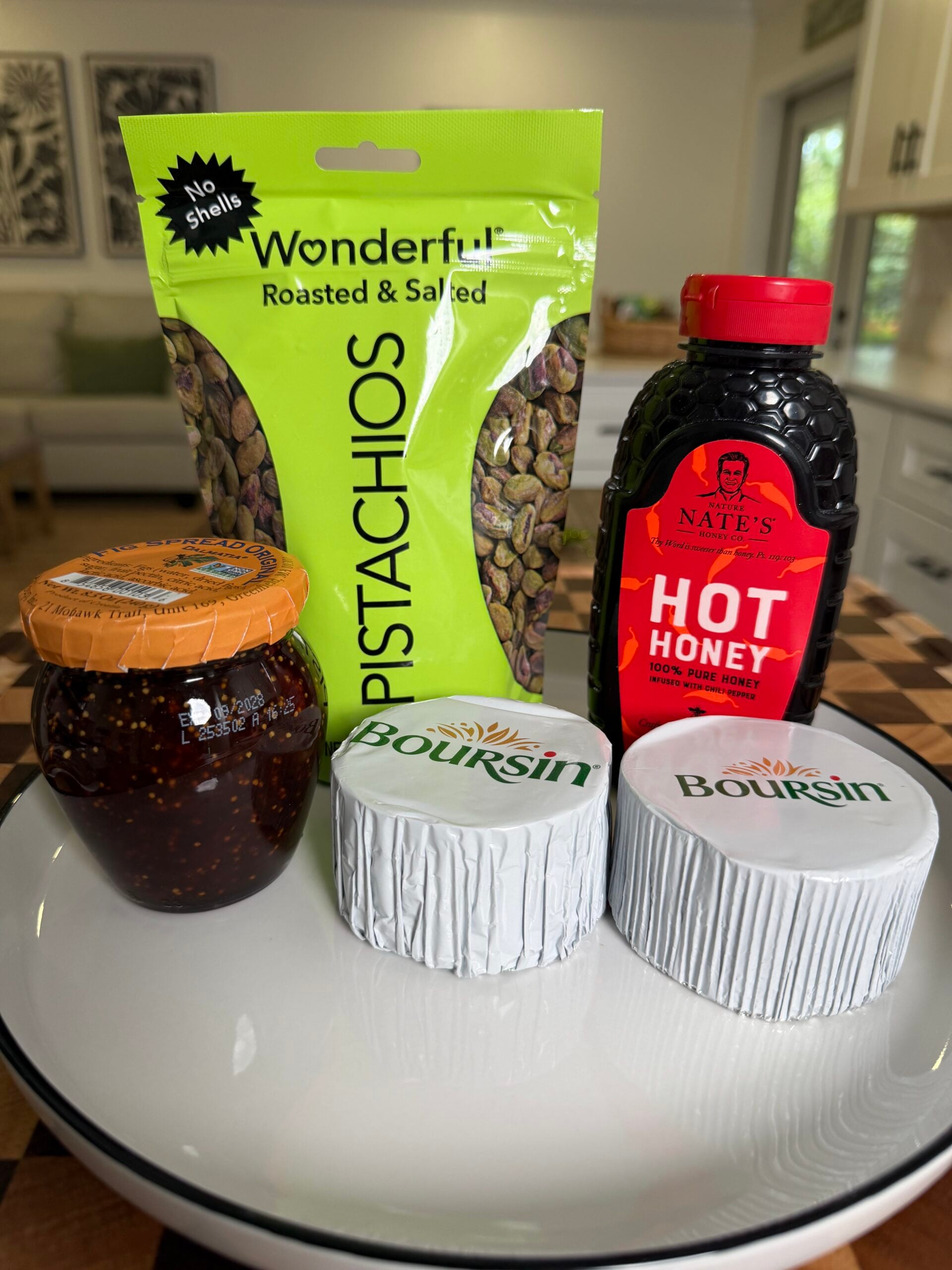 A bag of Wonderful roasted and salted pistachios, a bottle of Nates Hot Honey, a jar of fig spread, and two wheels of Boursin cheese on a plate.