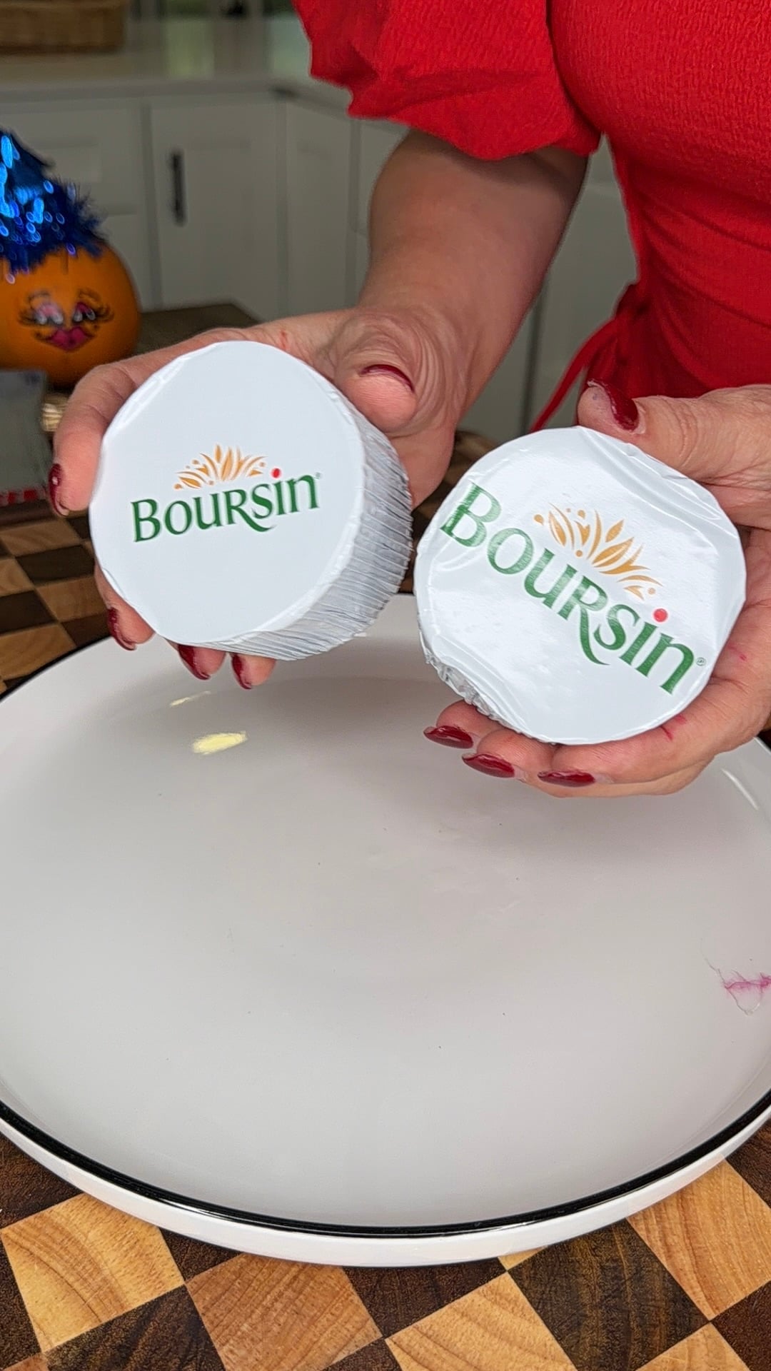 A person in a red shirt holds two unopened Boursin cheese rounds over a white plate.