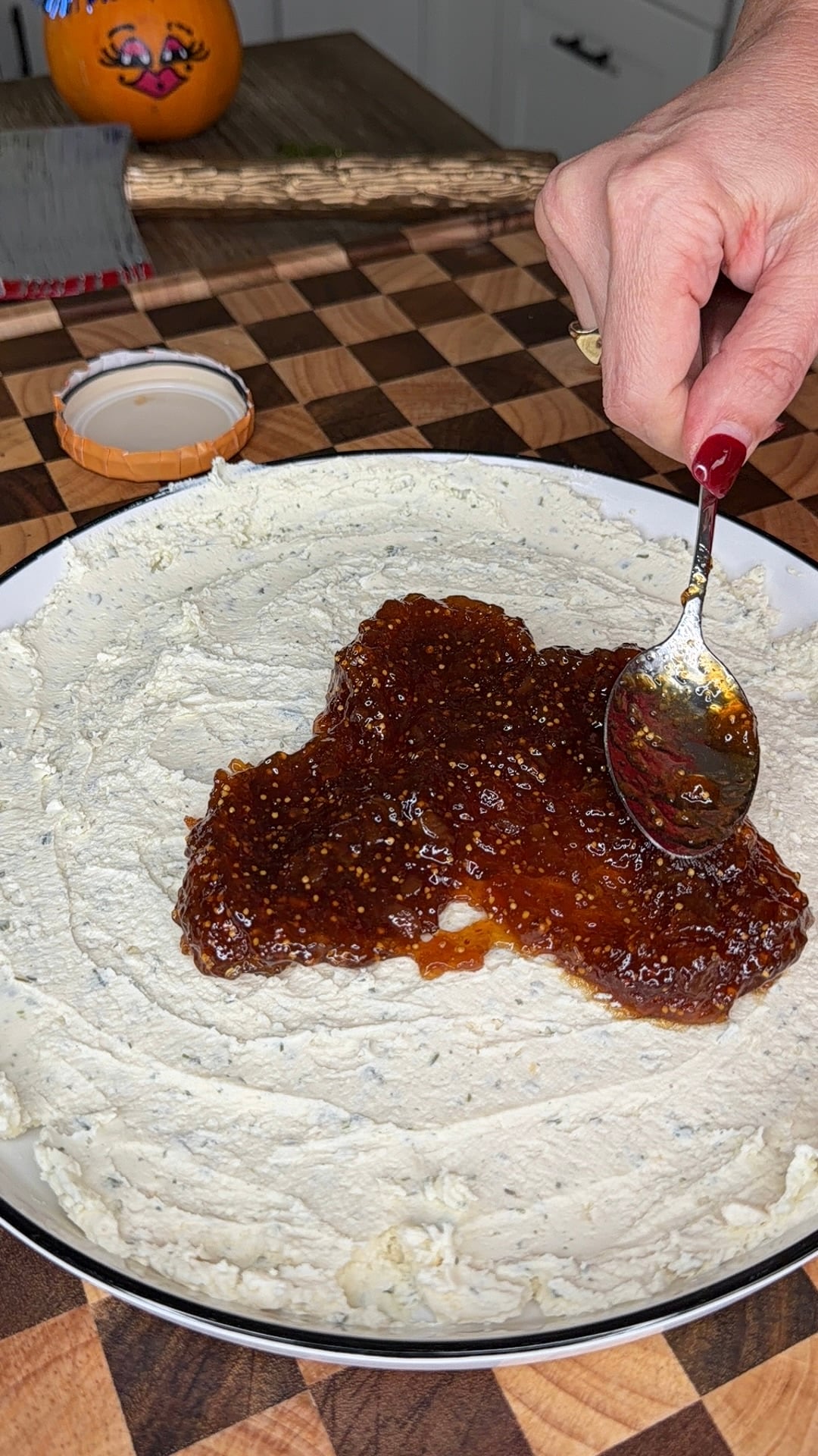 A hand uses a spoon to spread fig jam over a layer of cream cheese on a platter.