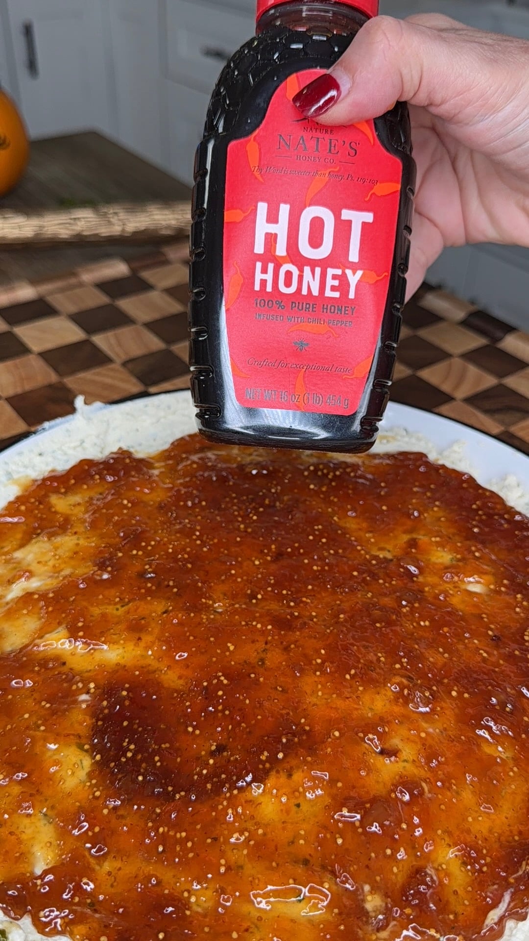 A hand squeezing a bottle of Nature Nates Hot Honey over a circular dish coated in a thick, reddish-brown sauce.