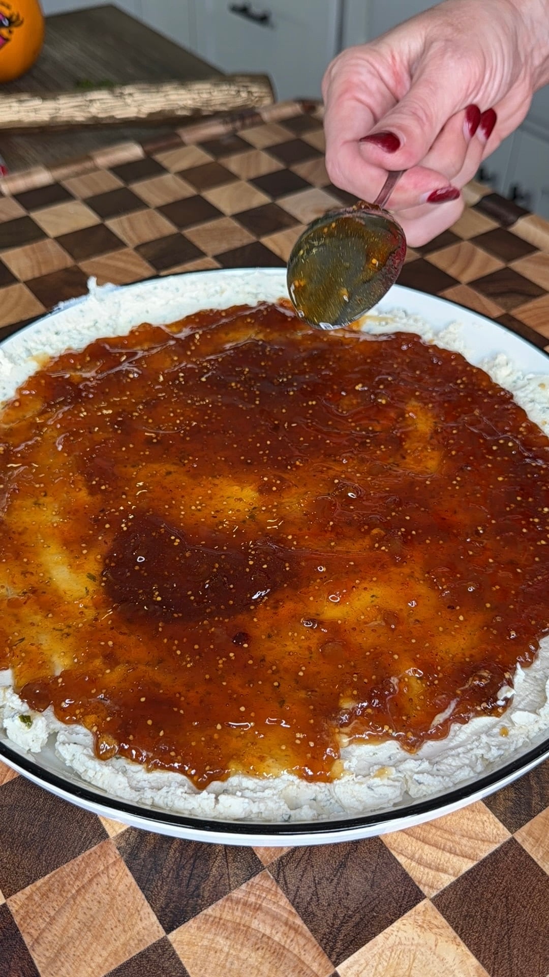 A hand uses a spoon to spread fig jam over a layer of soft white cheese in a shallow dish.