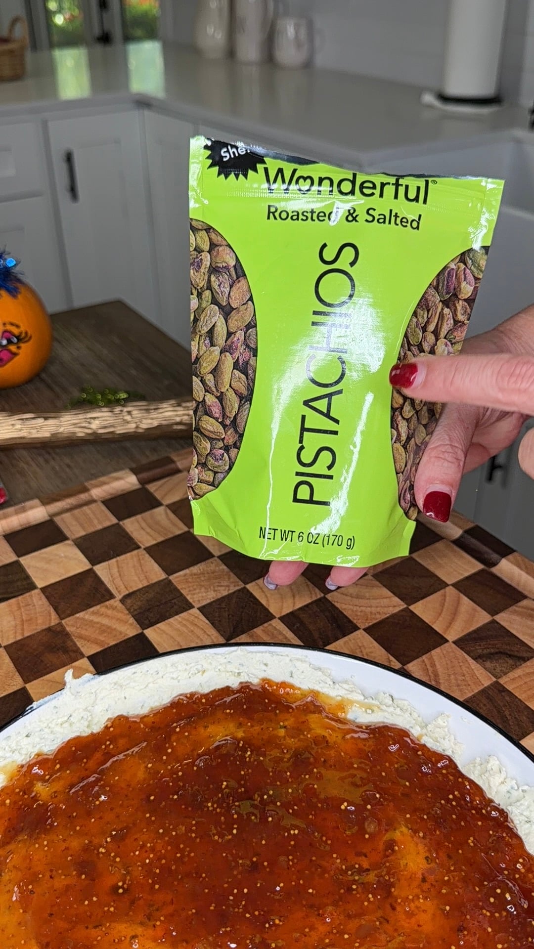 A hand holds a bag of Wonderful Roasted & Salted Pistachios over a round dish covered in cream cheese and a spread of fig jam.