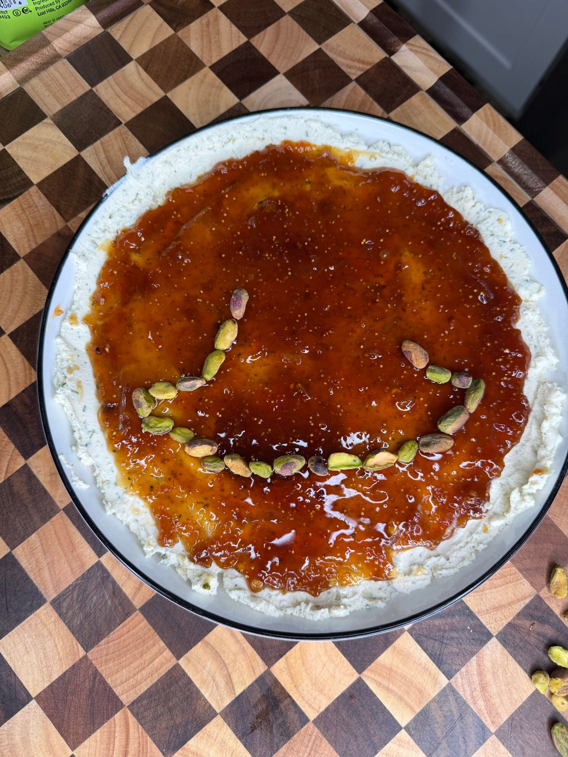A round serving plate of cream cheese topped with fig jam and a smiley face made of shelled pistachios.