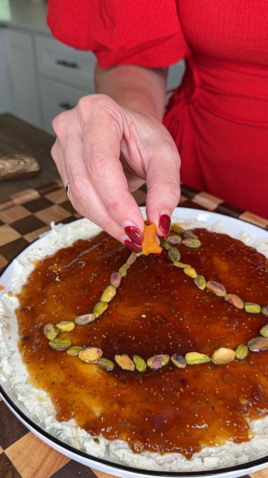 A person with red nail polish uses a small cracker to garnish a plate of spread topped with fig jam and a triangle of pistachios.