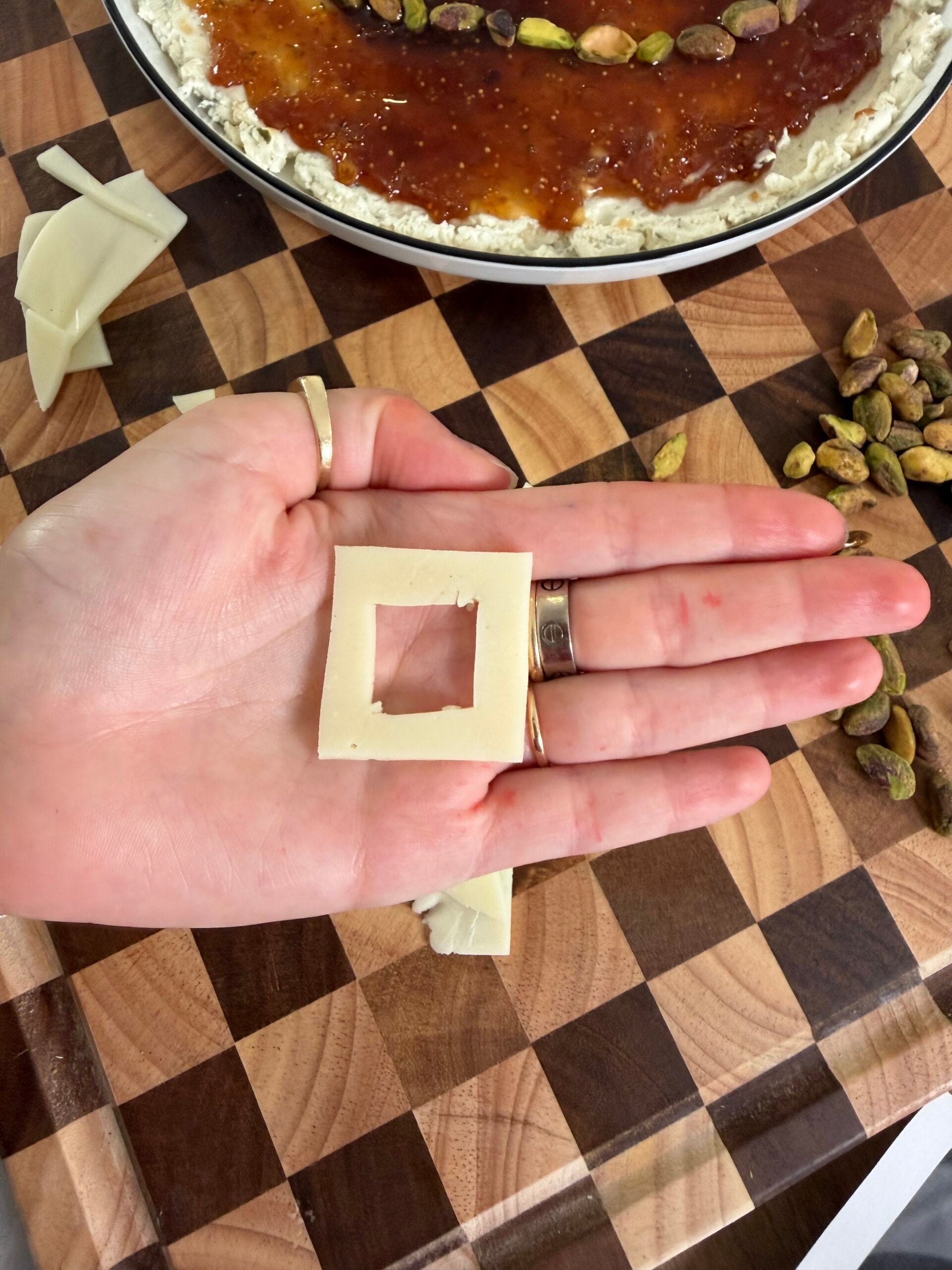 A hand holding a square slice of cheese with a square hole cut out of its center, over a checkered wooden cutting board with pistachios and a spread-topped appetizer.