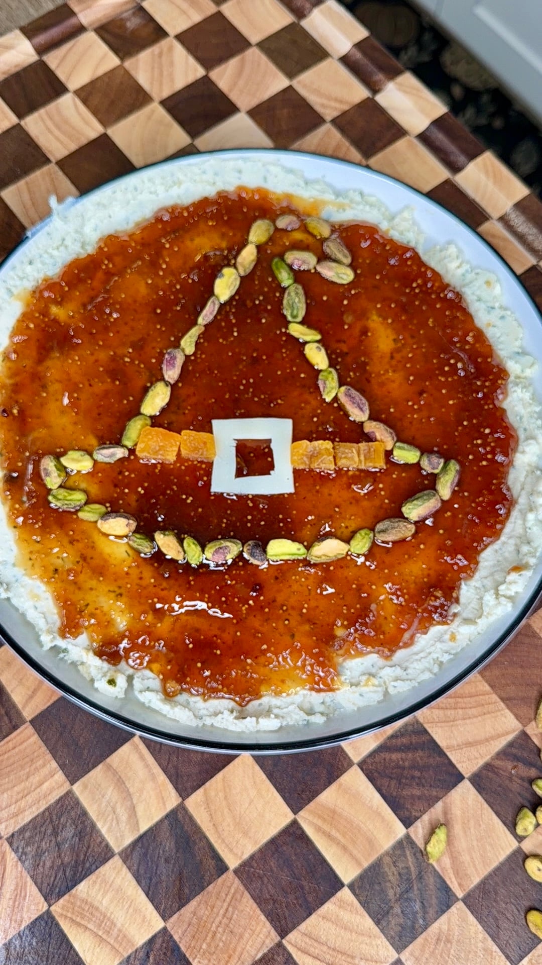 A round appetizer spread on a platter, topped with a layer of fig jam decorated with pistachios and dried fruit to resemble a witchs hat.