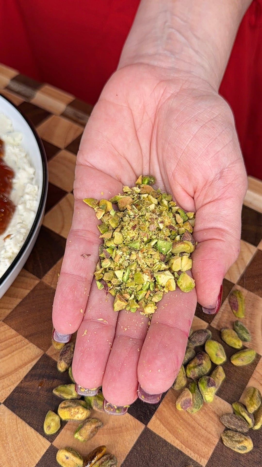 A persons hand holds a pile of chopped green pistachios over a checkered cutting board.