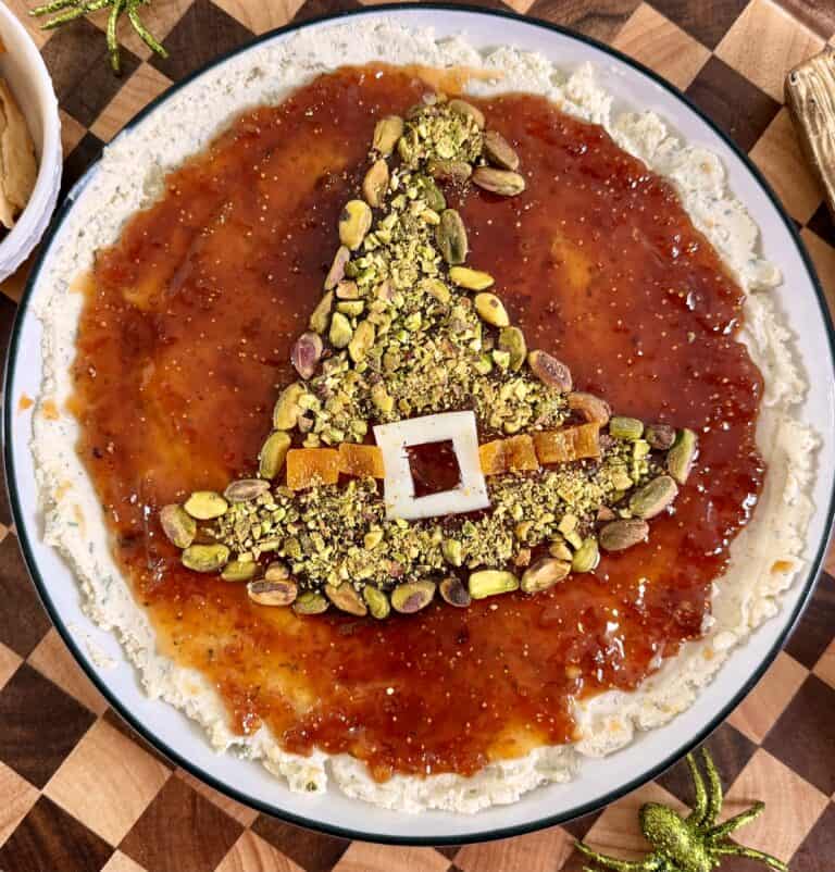 A round appetizer plate featuring a witchs hat shape made of crushed pistachios and dried fruit on a base of red jam and cream cheese.