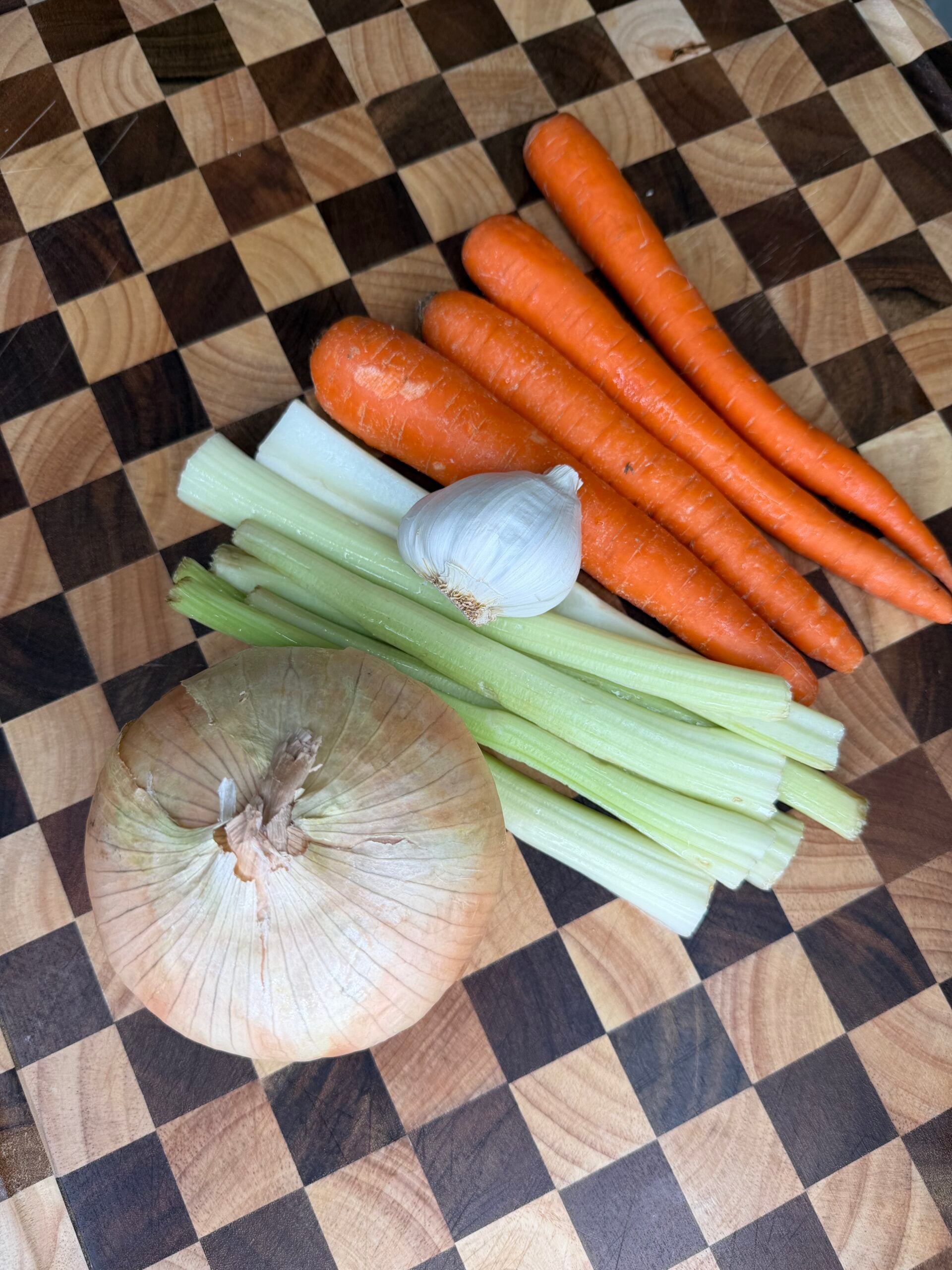 A whole onion, a bulb of garlic, several stalks of celery, and four carrots arranged on a checkerboard cutting board.
