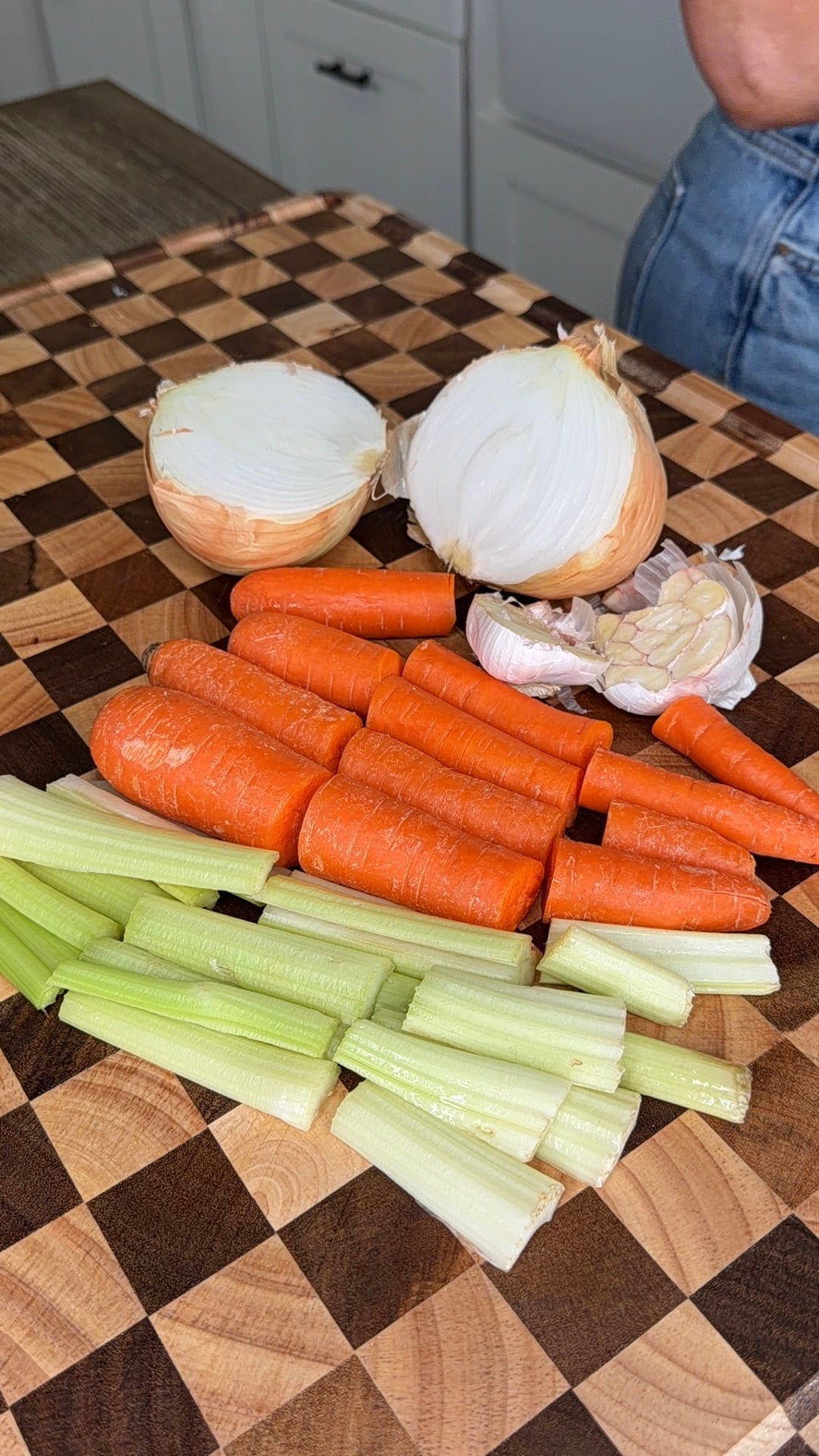 A wooden cutting board with chopped carrots, celery stalks, a halved onion, and a head of garlic.