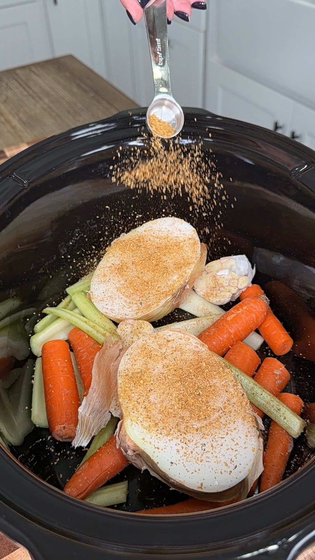 A hand holding a measuring spoon sprinkles seasoning over carrots, celery, and onion halves inside a slow cooker.