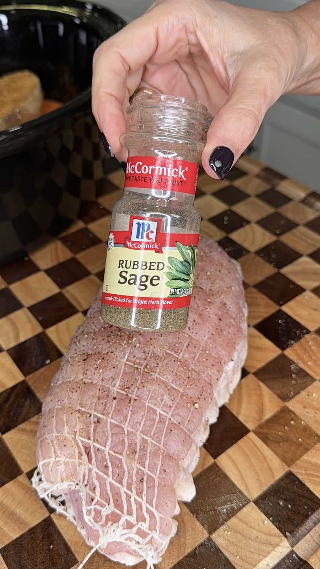 A hand holding a spice jar of McCormick Rubbed Sage over a raw, netted turkey breast on a checkered cutting board.