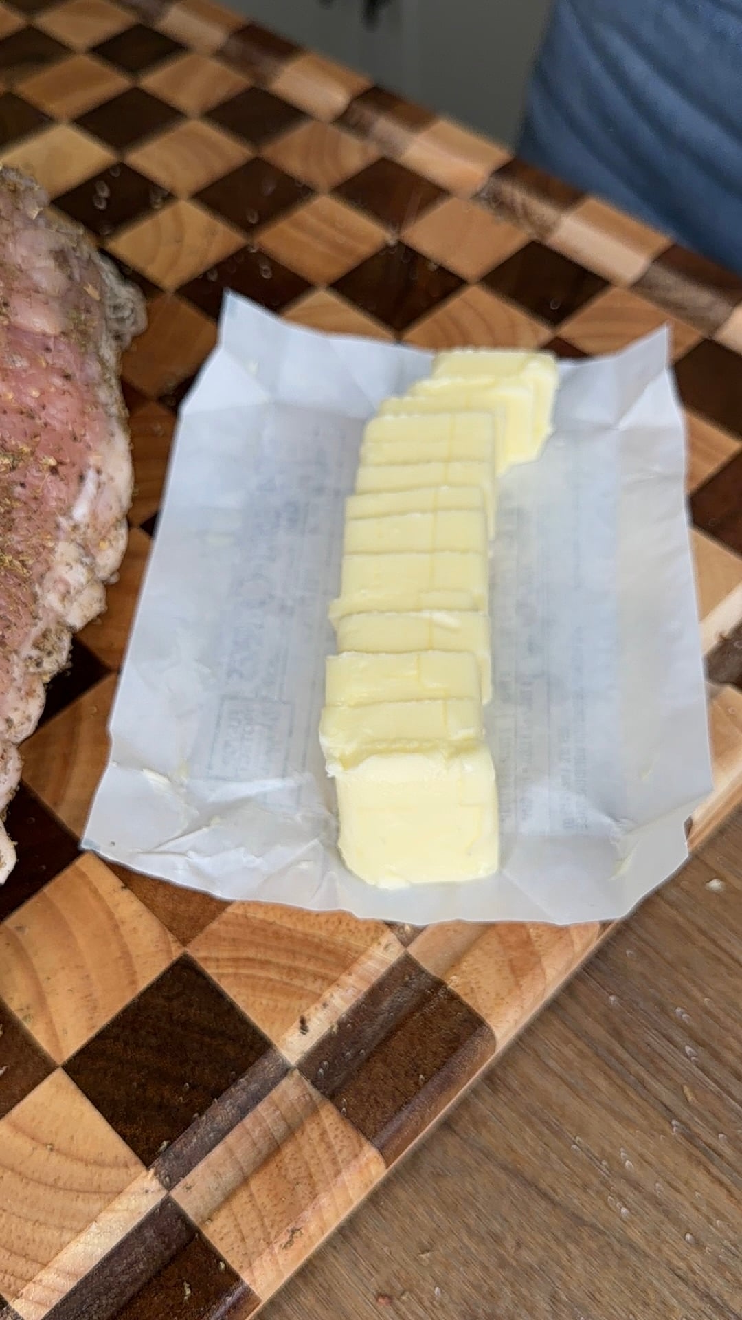 A stick of butter sliced into pats on a piece of parchment paper next to a seasoned piece of meat on a checkered cutting board.