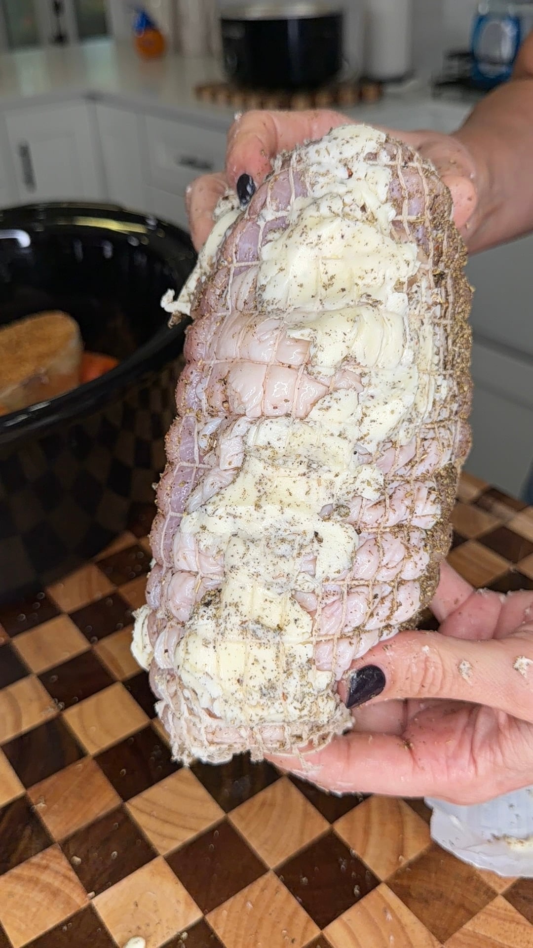 A person holds a netted raw roast seasoned with butter and herbs over a checkered cutting board.