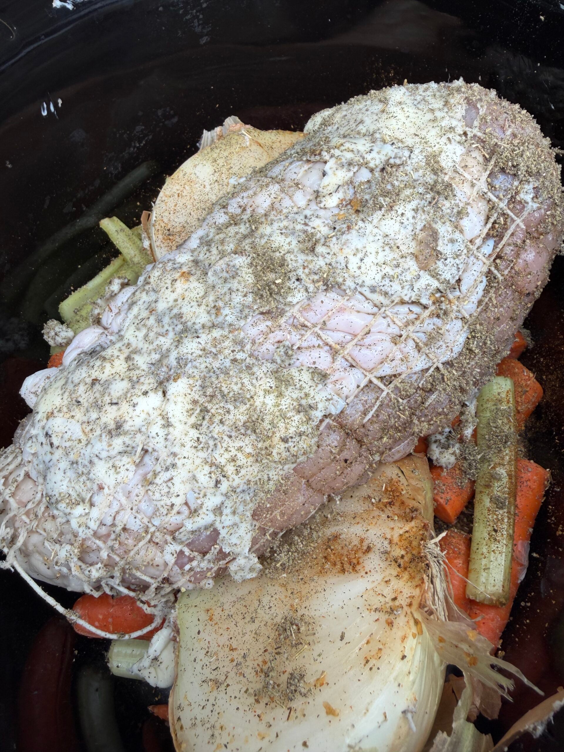 A seasoned, netted roast sits in a slow cooker atop chopped carrots, celery, and onion halves.