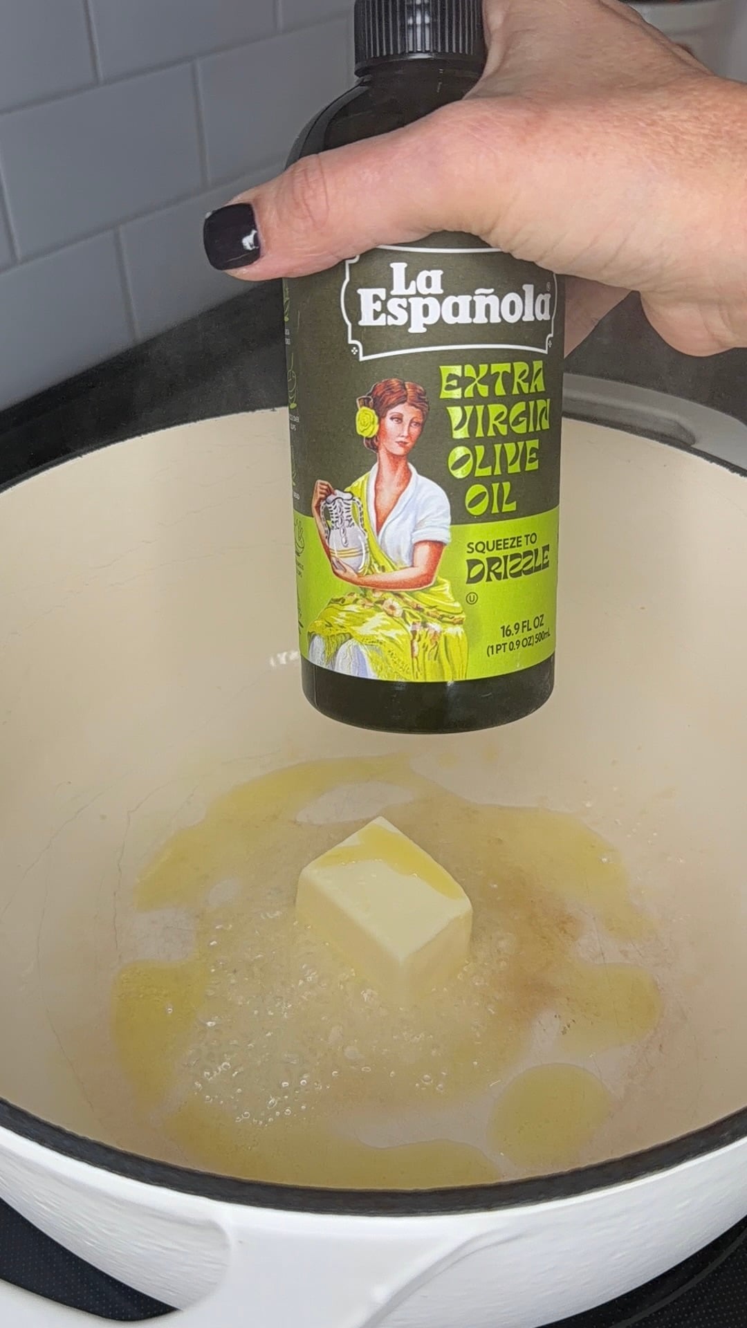 A hand holding a bottle of La Española extra virgin olive oil over a pat of melting butter in a Dutch oven.