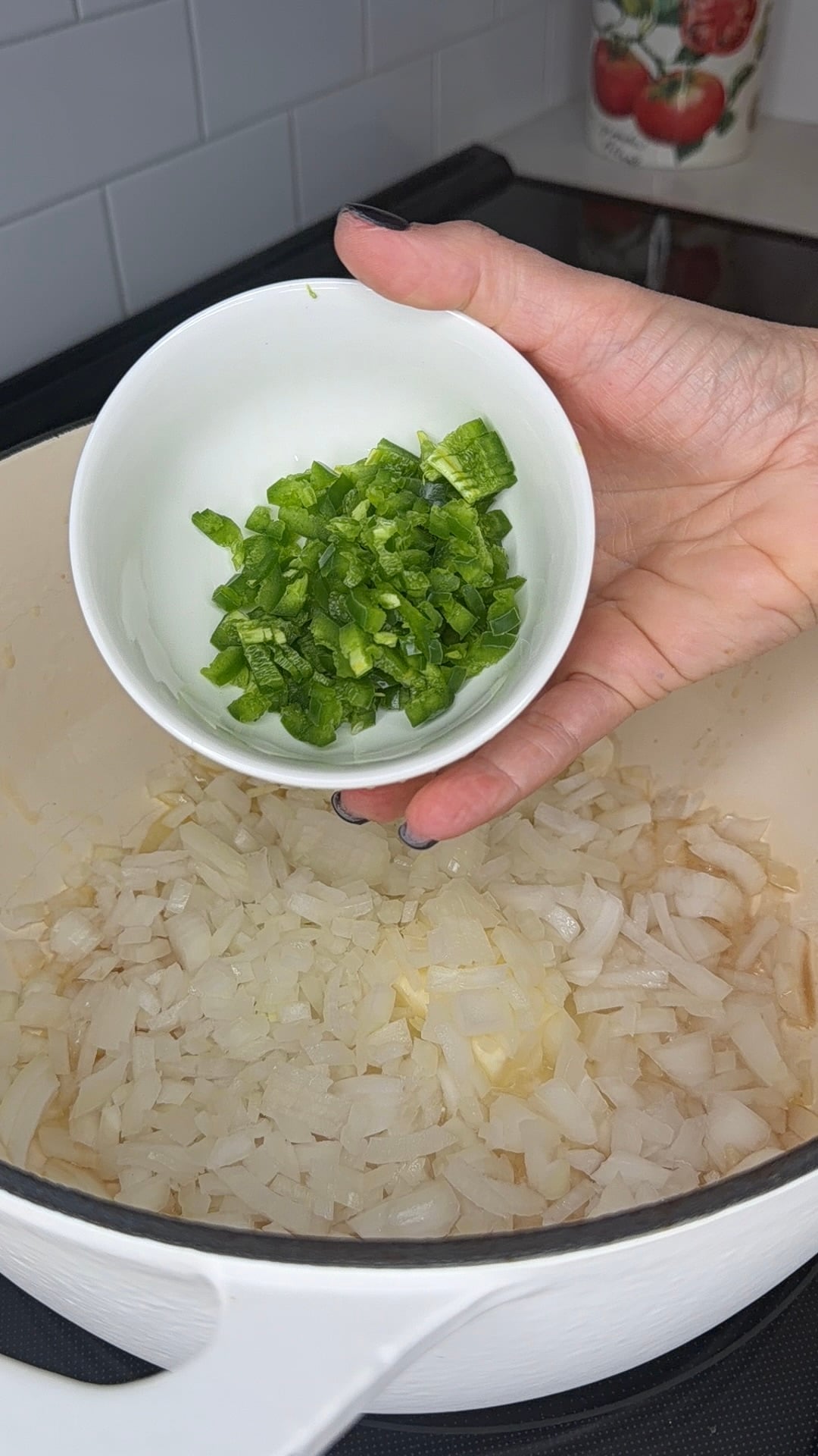 A hand holds a small white bowl of diced green jalapeños over a pot containing chopped sautéing onions.