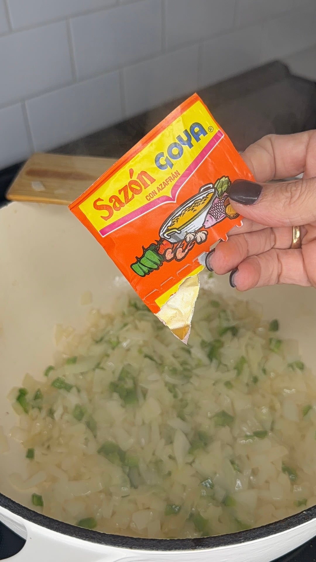 A hand pours a packet of Sazón Goya con Azafrán seasoning into a pot of sautéing onions and peppers.