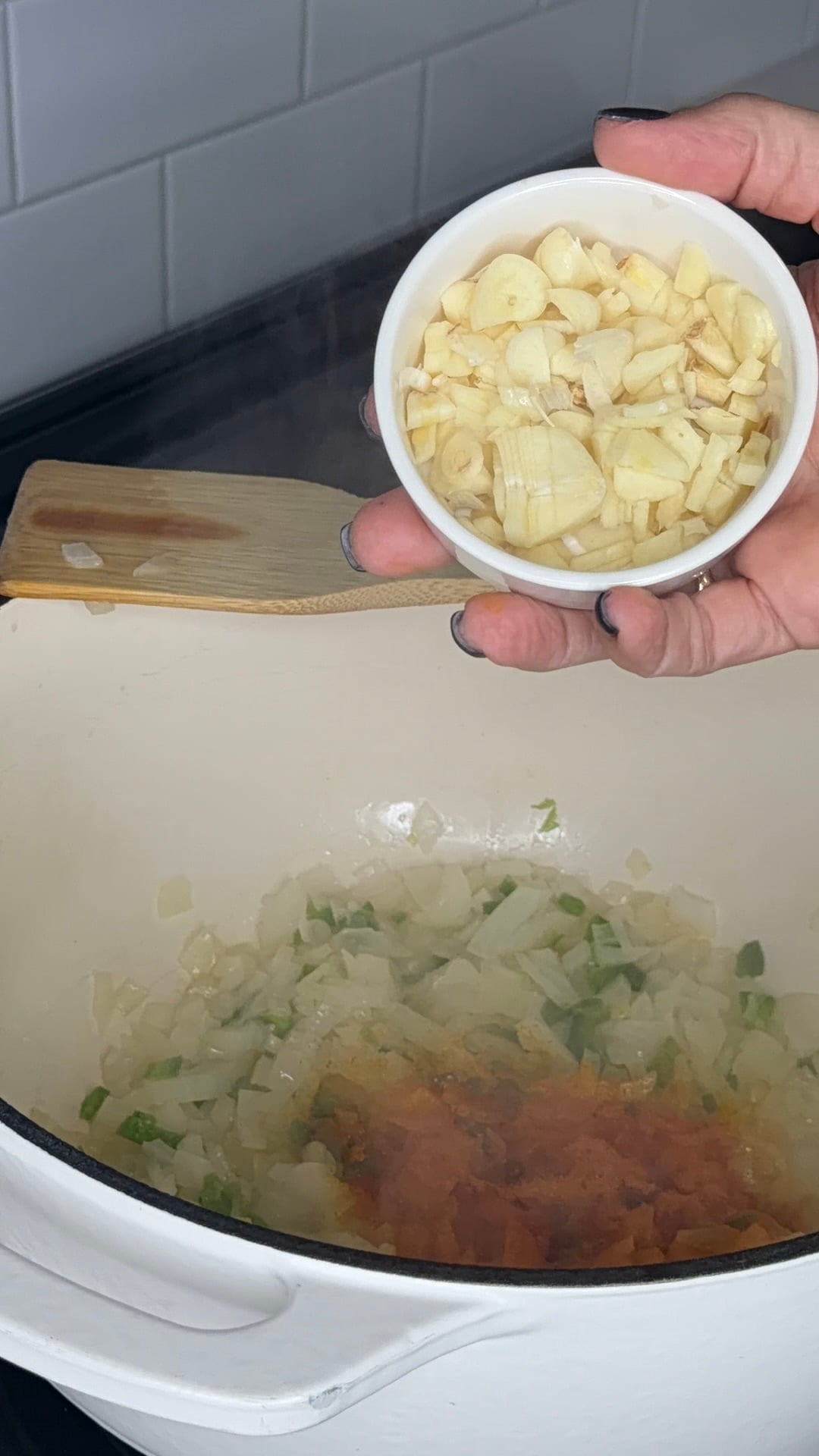 A hand holds a small bowl of chopped garlic over a pot containing sautéed onions, peppers, and tomato paste.