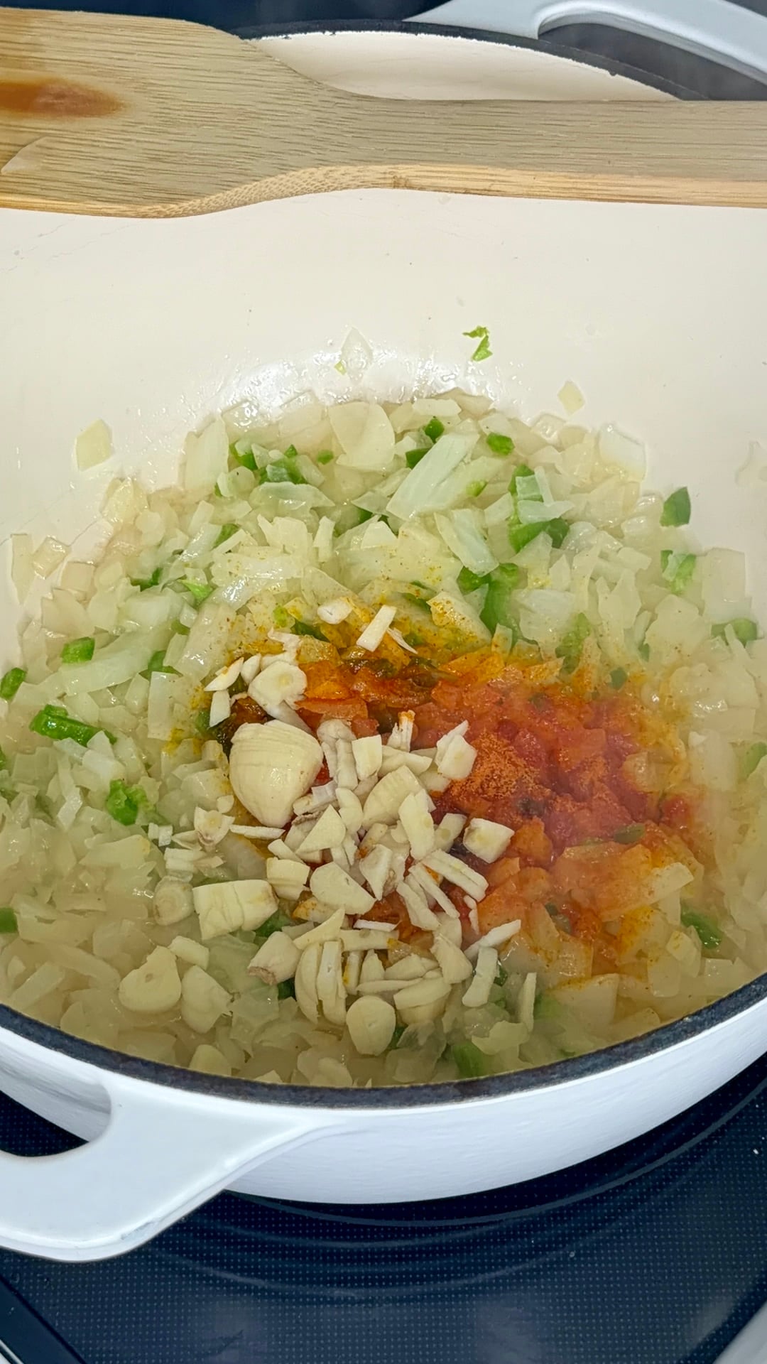 Chopped onions, green peppers, garlic, and spices cooking in a white pot with a wooden spoon resting on top.