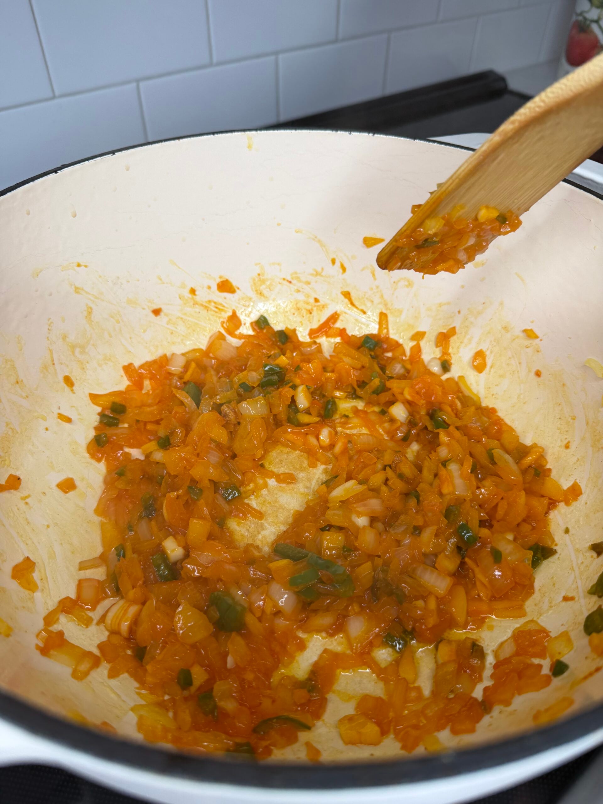 A wooden spoon stirring a mixture of sautéed diced carrots, onions, and green herbs in a large enamel pot.