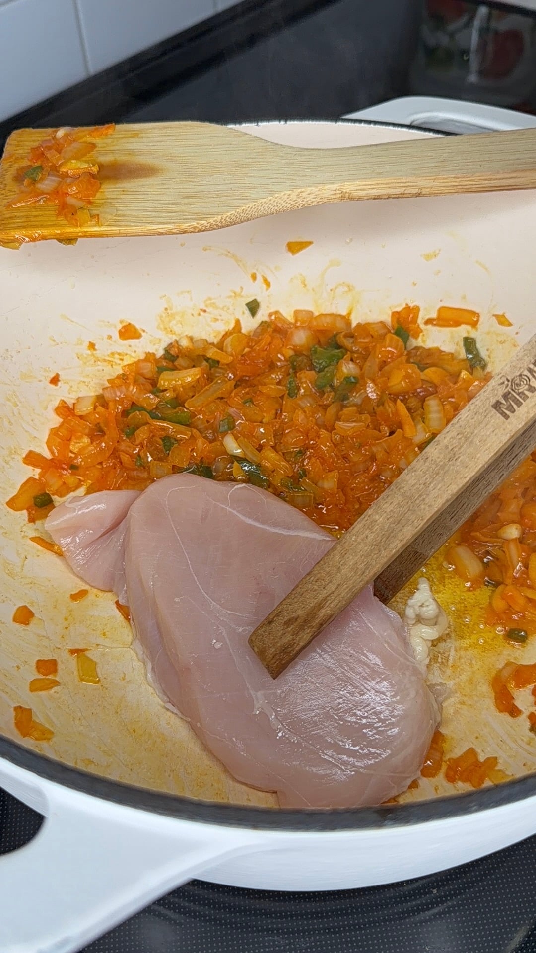 A raw chicken breast being pressed into a pan with sautéed onions and peppers by a wooden spatula.