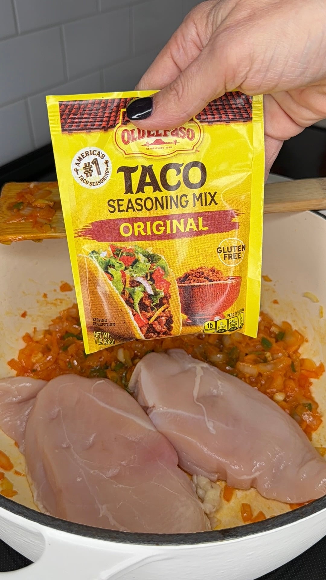 A hand holds a packet of Old El Paso Original Taco Seasoning Mix over a pot containing raw chicken breasts and sautéed vegetables.