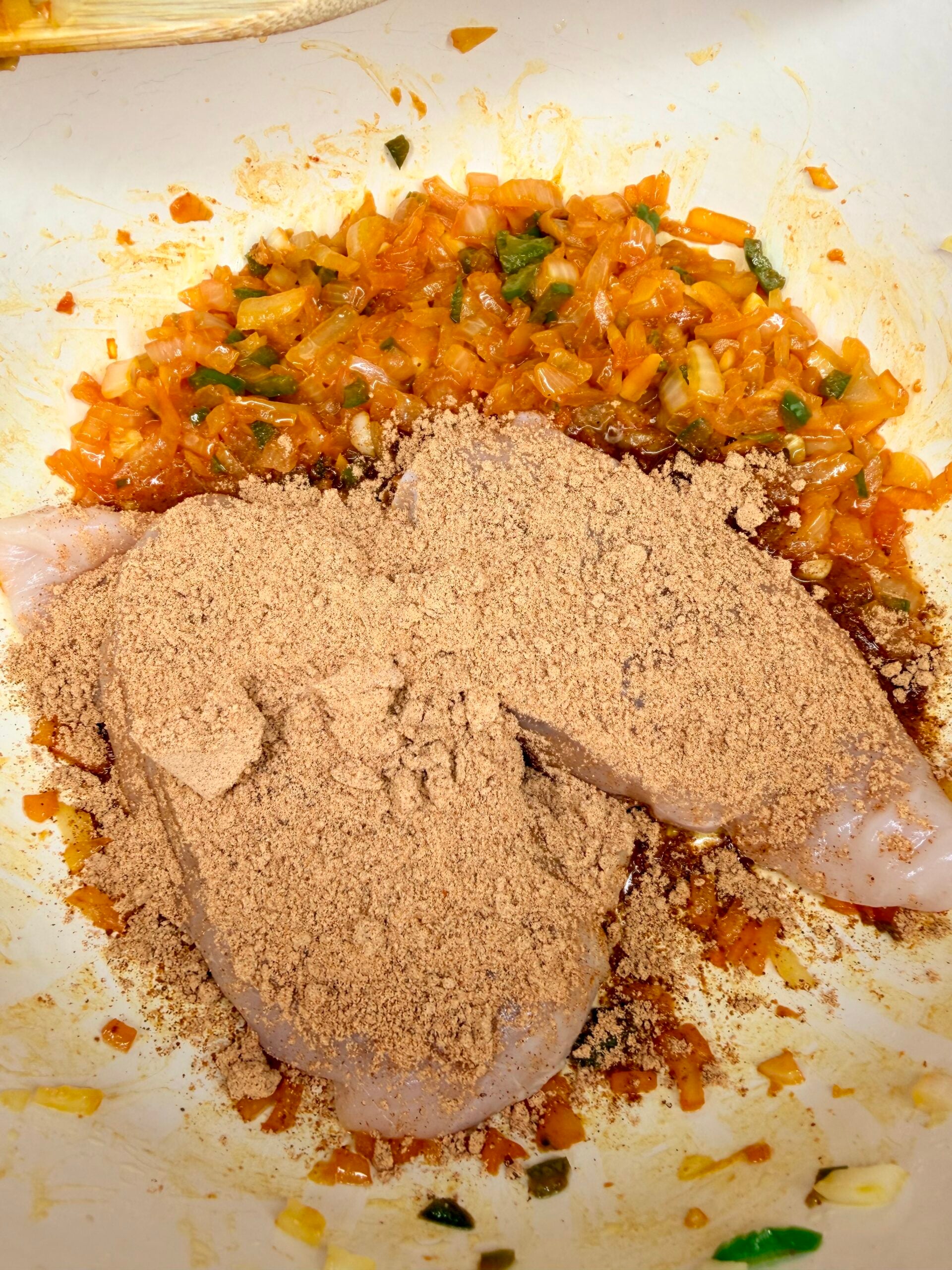 Raw chicken breast coated in a thick layer of brown seasoning, sitting in a pan with sautéed onions and peppers.